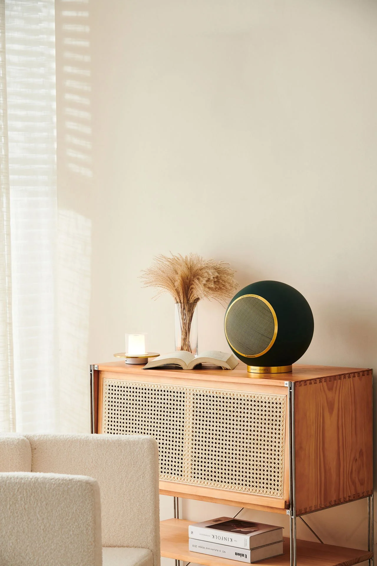 Ein Holzregal mit einer Rattan-Tür, darauf befinden sich eine Glast Schale mit Gräsern, ein Buch, ein Lautsprecher, eine kleine Lampe und unten zwei Bücher.