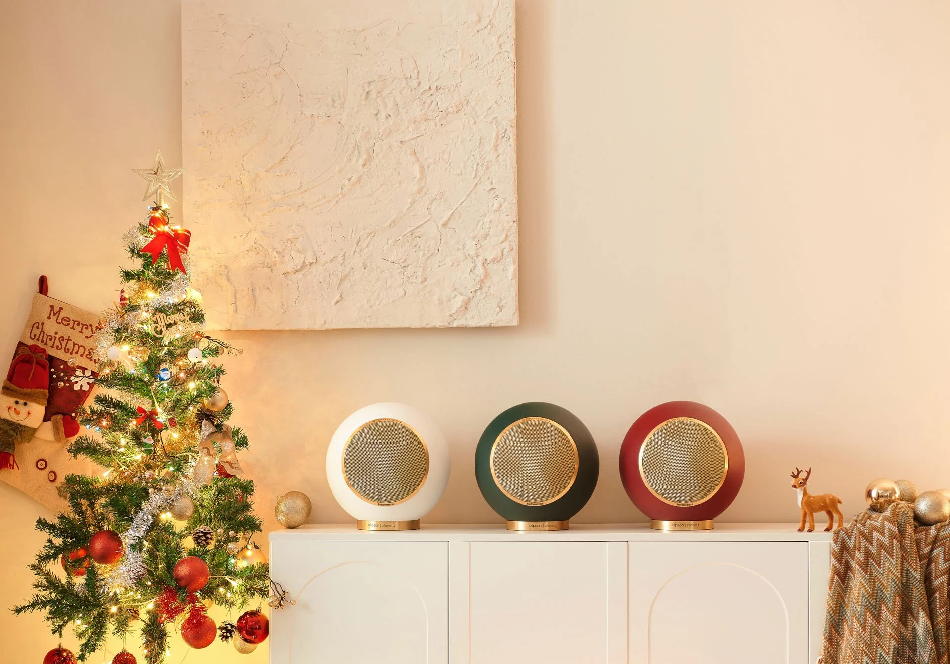 Weihnachtsschmuck in einem Wohnzimmer, Weihnachtsbaum mit Kugeln, Lichter und Stern, auf einer weißen Kommode drei Lautsprecher in weiß, grün und rot, kleine Deko-Figur eines Rehs, goldene Weihnachtskugeln, weihnachtlicher Stoff