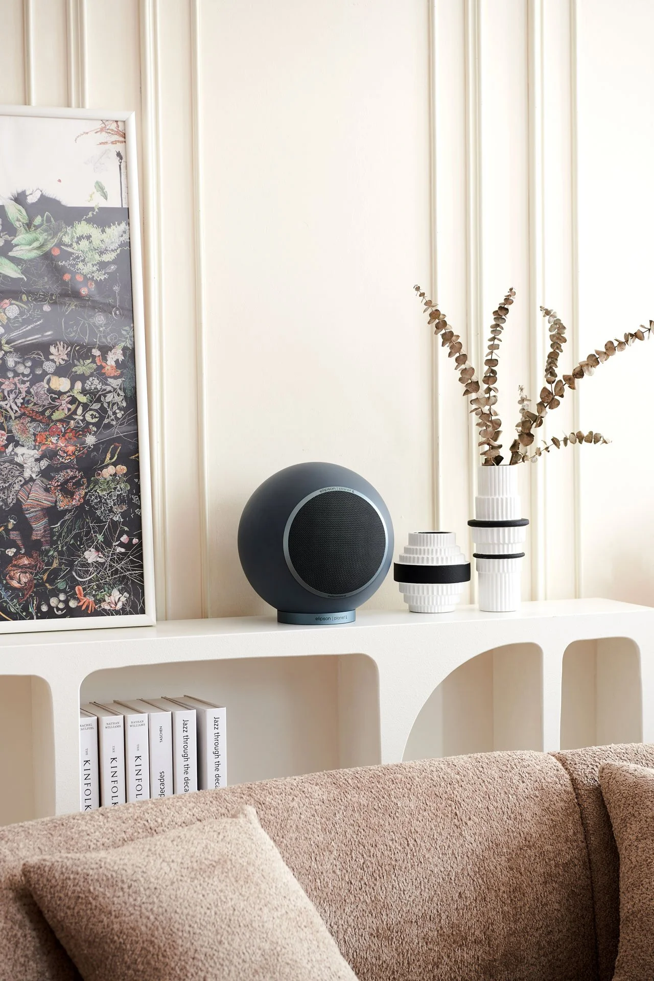 Wohnzimmer mit weißen Wandpaneelen, einem weißen Regal mit Büchern, einer braunen Couch mit Kissen, einem schwarzen Lautsprecher, und weißen Vasen mit getrockneten Zweigen.