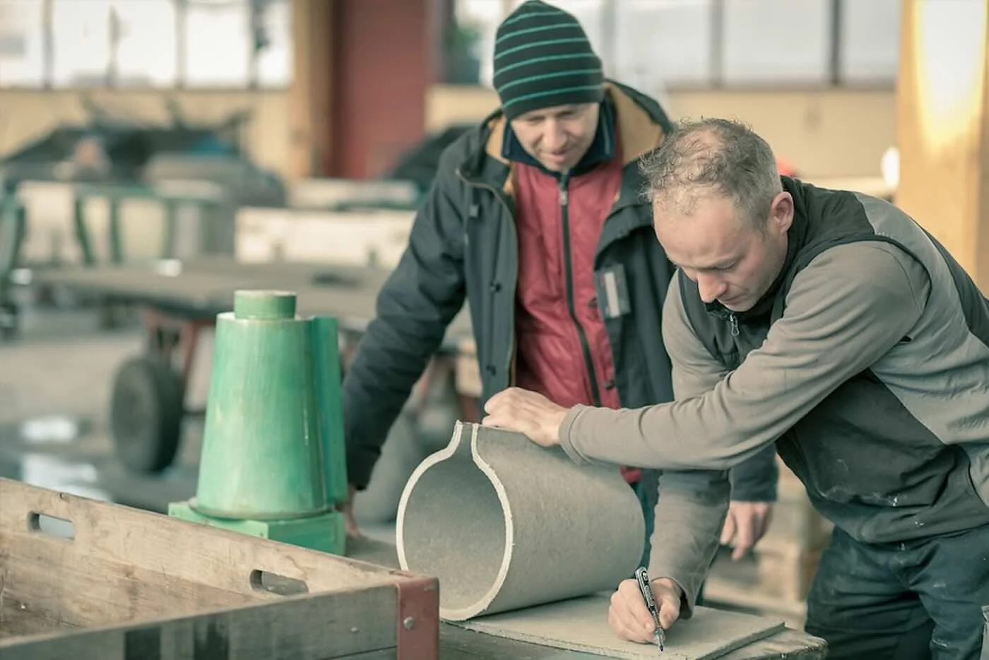 Zwei Männer arbeiten in einer Werkstatt an einem großen, runden Gegenstand aus Stein. Einer der Männer schreibt auf das Material, während der andere zuschaut.
