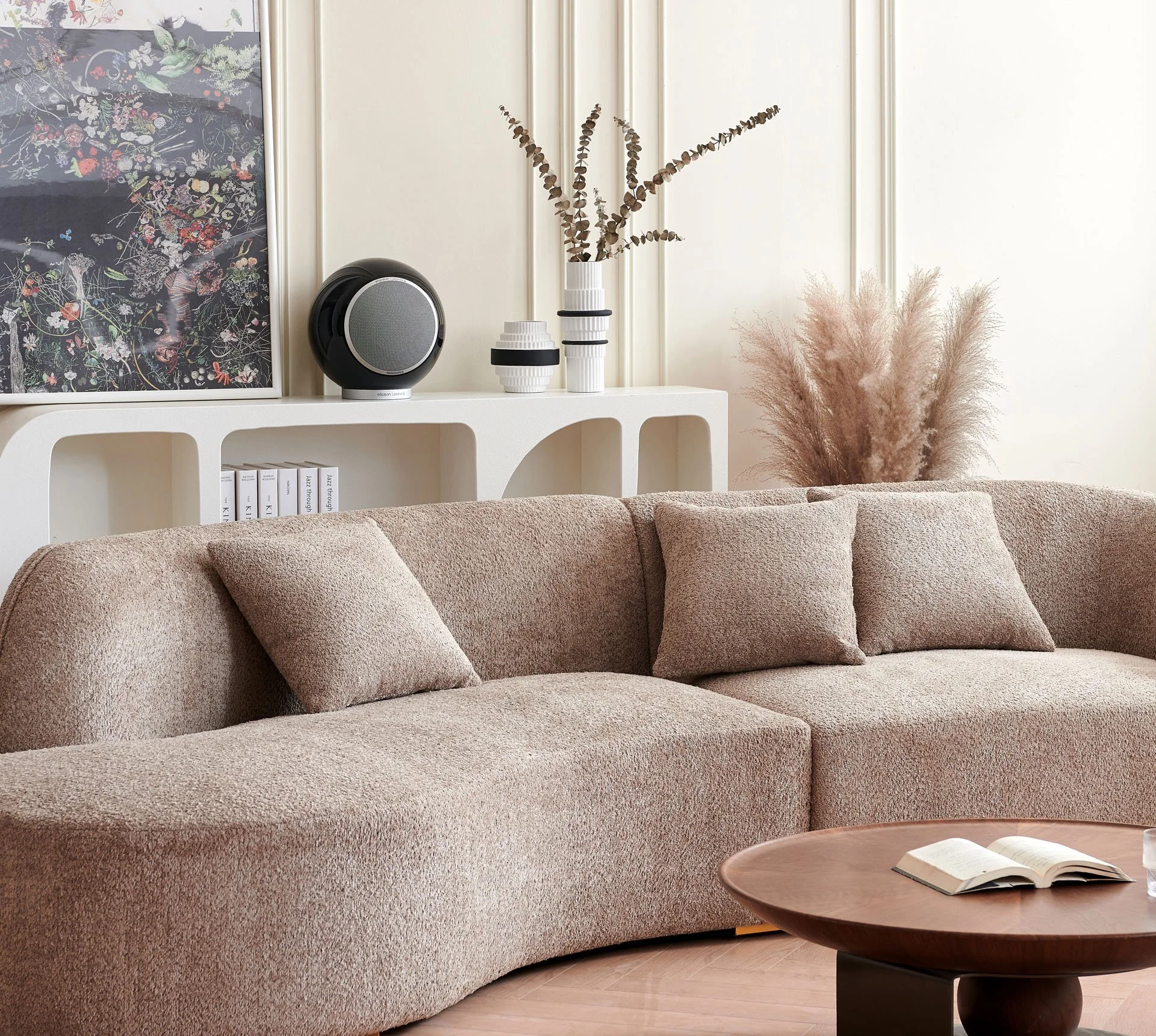 Wohnzimmer mit einer beige Couch, einem runden Holztisch, weißen Wandregalen mit dekorativen Vasen, Büchern und einem Lautsprecher. Es gibt auch eine Wandzeichnung und Pflanzen, darunter pampas Gras.