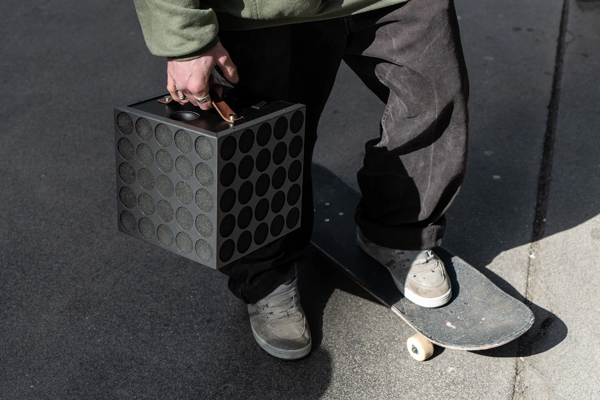 Person mit Skateboard, hält einen schwarzen Kasten mit Löchern, trägt beiges Schuhwerk, die Person steht auf Asphalt.