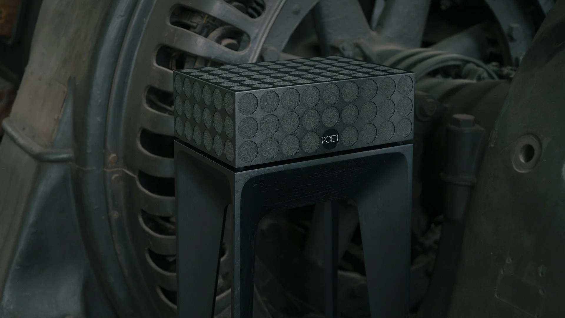 Ein schwarzer Lautsprecher mit runden Mustern auf dem Gehäuse, auf einem schwarzen Holzhocker vor einer unordentlichen Autowaschanlage