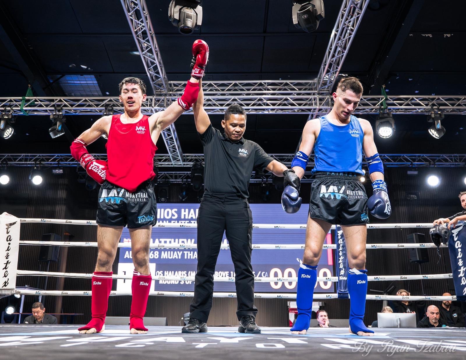 Two Muay Thai fighters in red and blue trunks standing with their hands raised by the referee inside a boxing ring. The referee is holding their hands up in victory, and the fighters are wearing gloves, shin guards, and mouthguards. The background shows an electronic scoreboard and lighting rig.