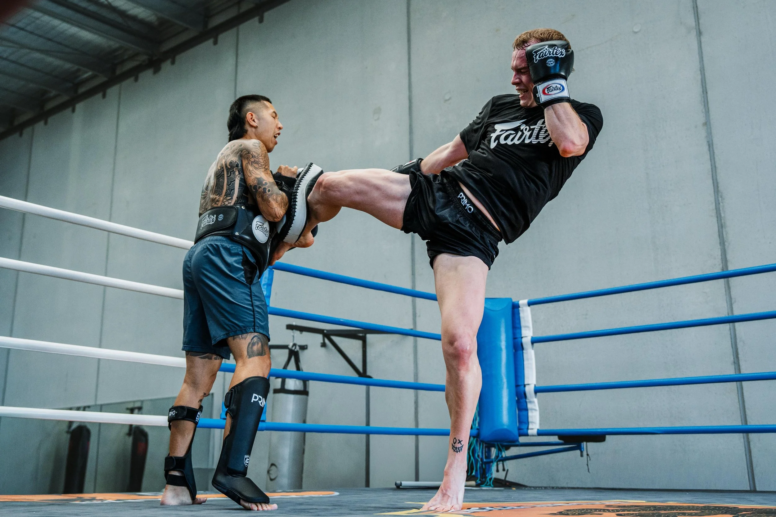 Two men engaged in a martial arts training session in a boxing ring inside a warehouse. One man, with tattoos, is preparing to kick the other man, who is wearing black gear and protective gloves. The man receiving the kick appears to be smiling or in a playful mood.