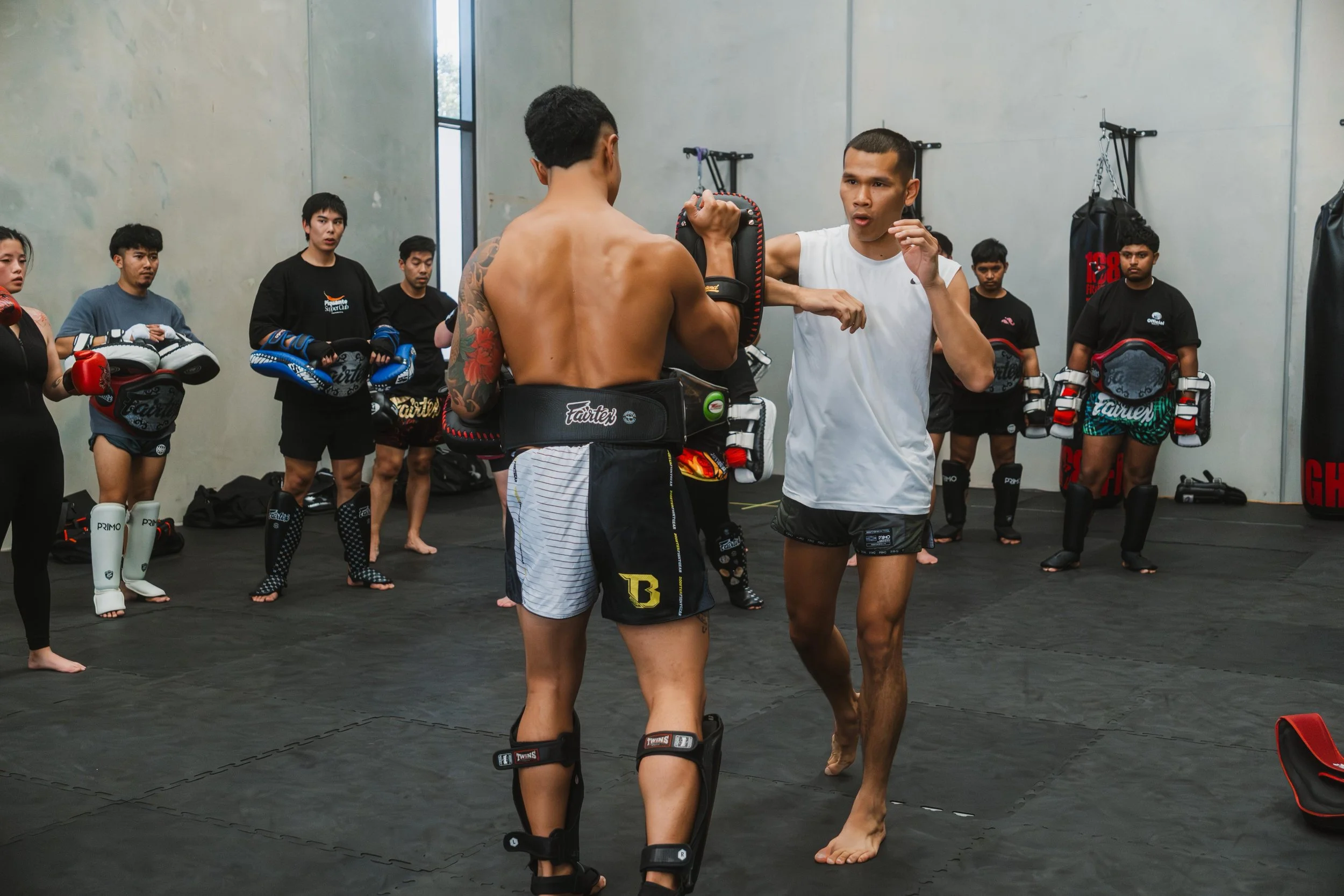 A martial arts or boxing class in session with students practicing with punching pads under the guidance of an instructor in a gym with gray walls and equipment hanging on the walls.