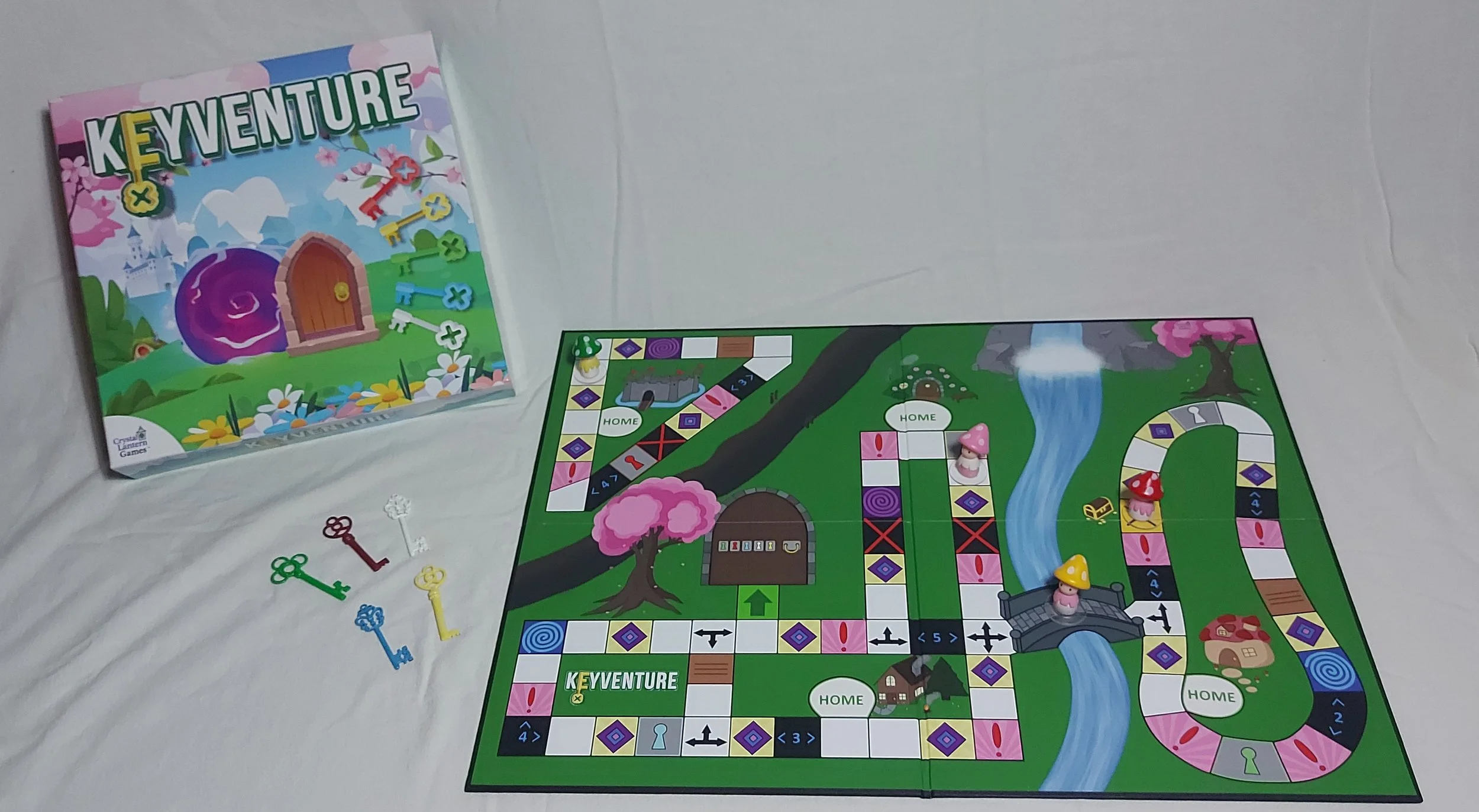 Board game called Keyventure with colorful game pieces, including red, white, green, yellow, and blue keys, and game board featuring paths, houses, a bridge, and a river, set on a white surface.