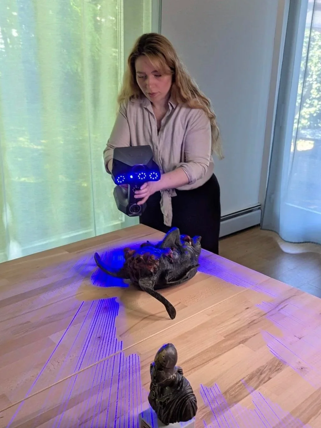 Frau mit 3D-Scanner, der eine Maske auf einem Tisch scannt, mit blauen Lichtern, im Raum neben Fenster und Vorhängen.