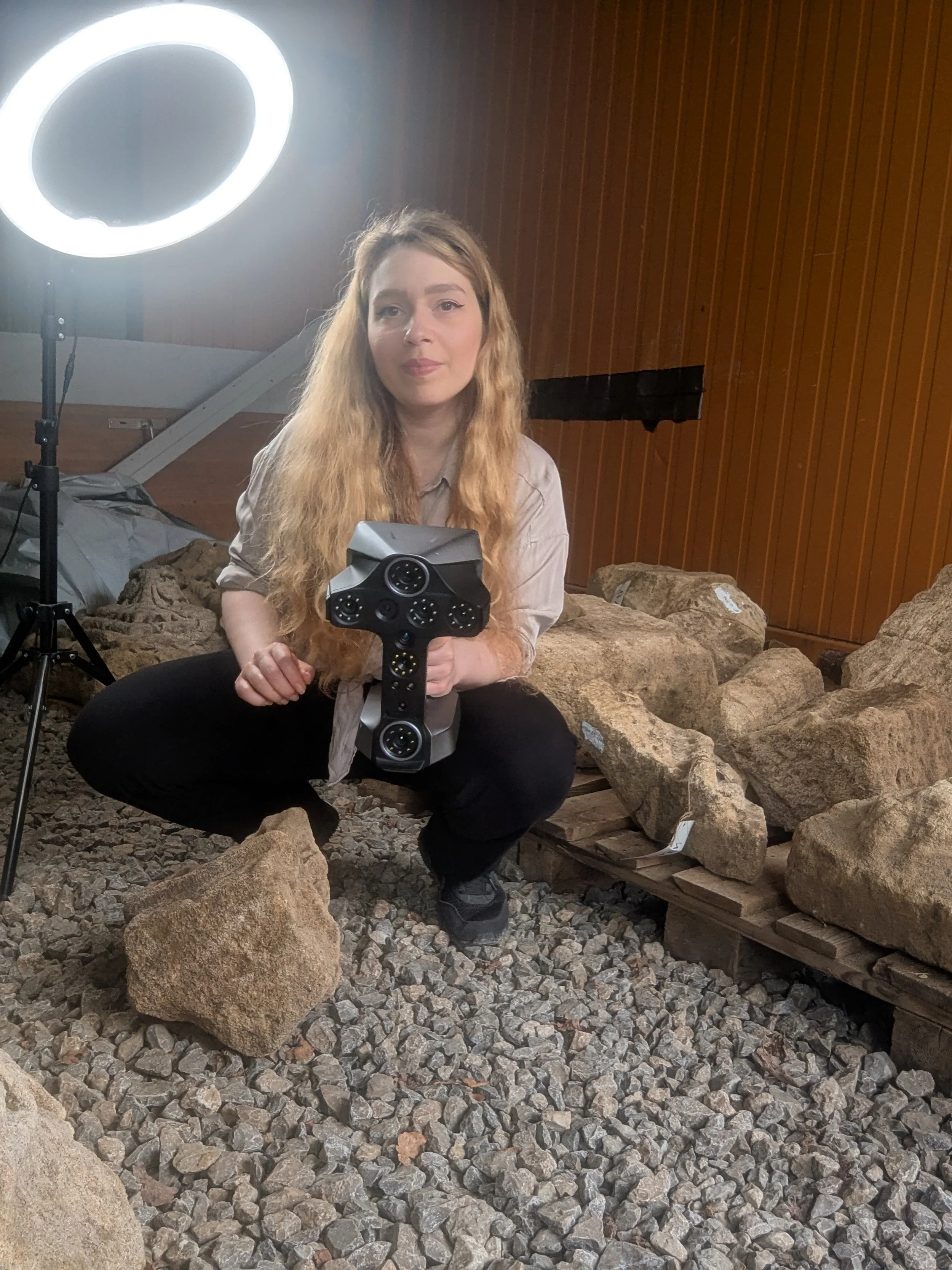 Eine junge Frau mit langen, lockigen, blonden Haaren hält einen 3D-Scanner in den Händen. Der Hintergrund besteht aus einer Holzwand und mehreren großen Steinen auf einem Kiesboden.