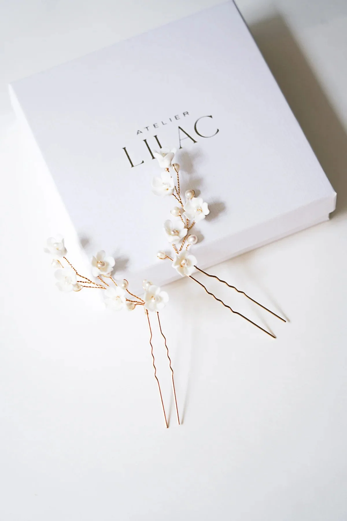 Deux épingles à cheveux décoratives avec des fleurs en perles blanches sur une boîte blanche portant l'inscription "Atelier Lilac".