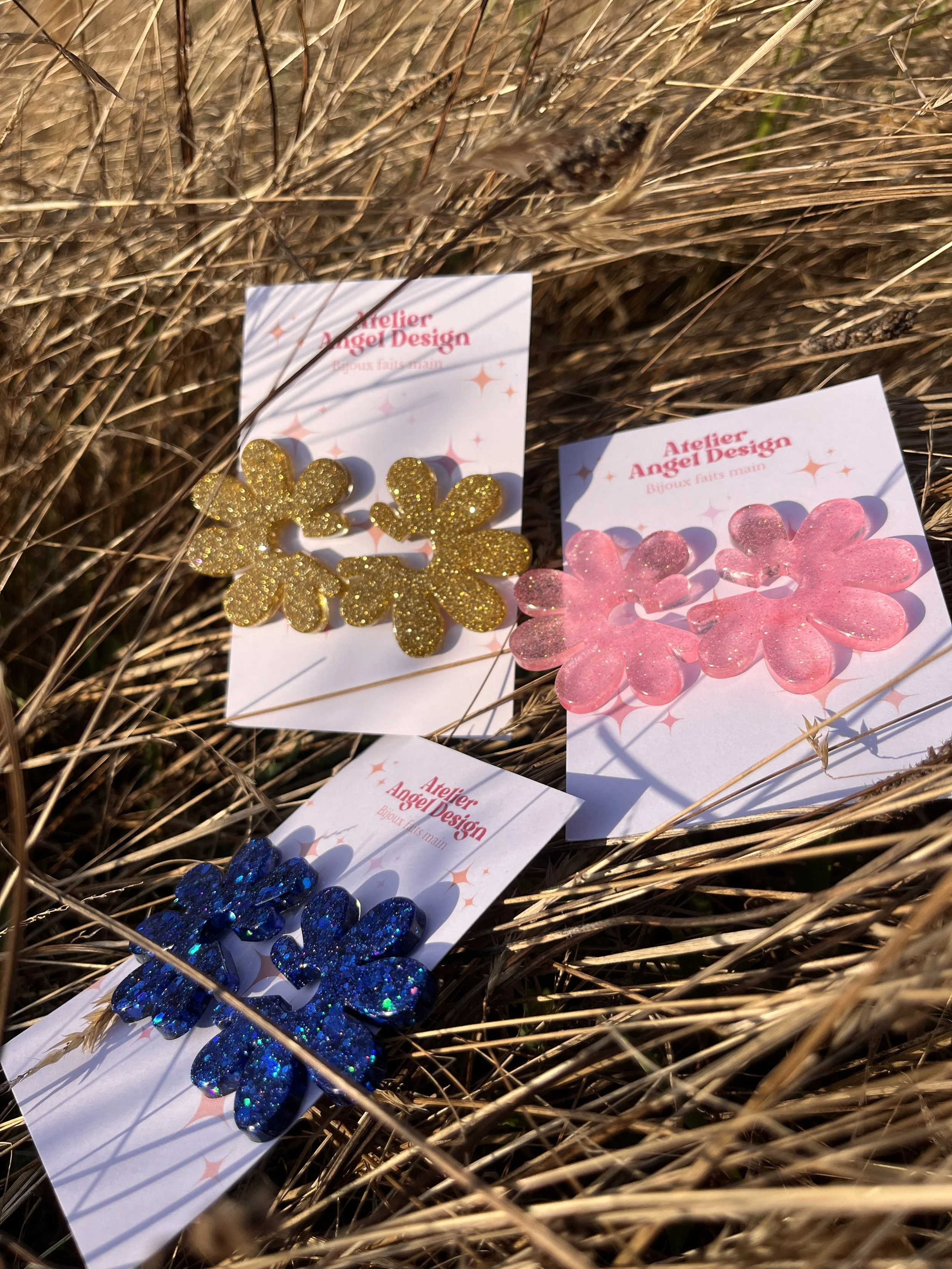 Trois paires de boucles d'oreilles en forme de marguerites avec paillettes, en or, rose et bleu, affichées sur des cartes de l'Atelier Angel Design, posées dans de l'herbe sèche.