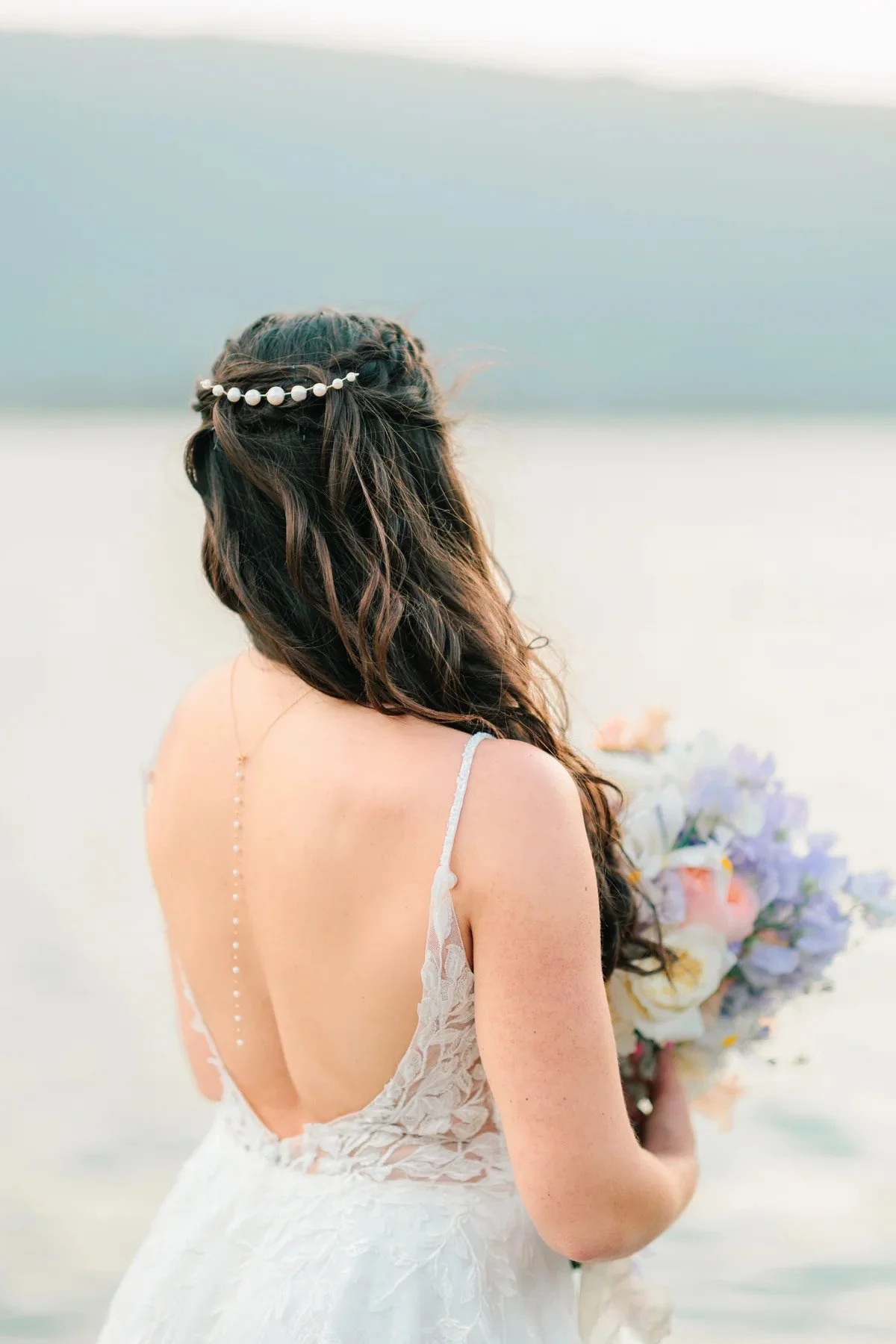 Femme en robe de mariée tenant un bouquet, vue de dos, sur une plage landaise avec dans les cheveux le peigne Lavande d'Atelier Lilac.