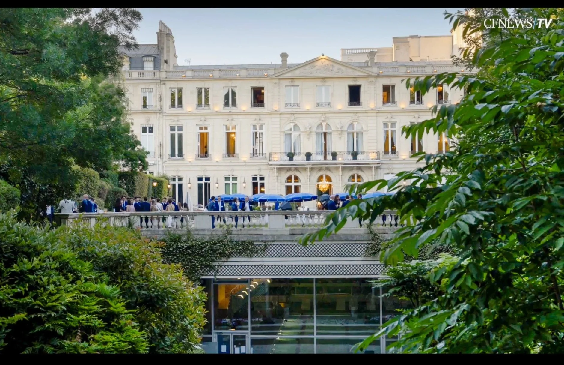 Un groupe de personnes en costume formel dans  jardin devant le cercle de l'union interralié. Arbres verts luxuriants, avec des parasols bleus au centre. L'image est prise en extérieur, lors d'une occasion ou d'un événement d'entreprise élégant.
