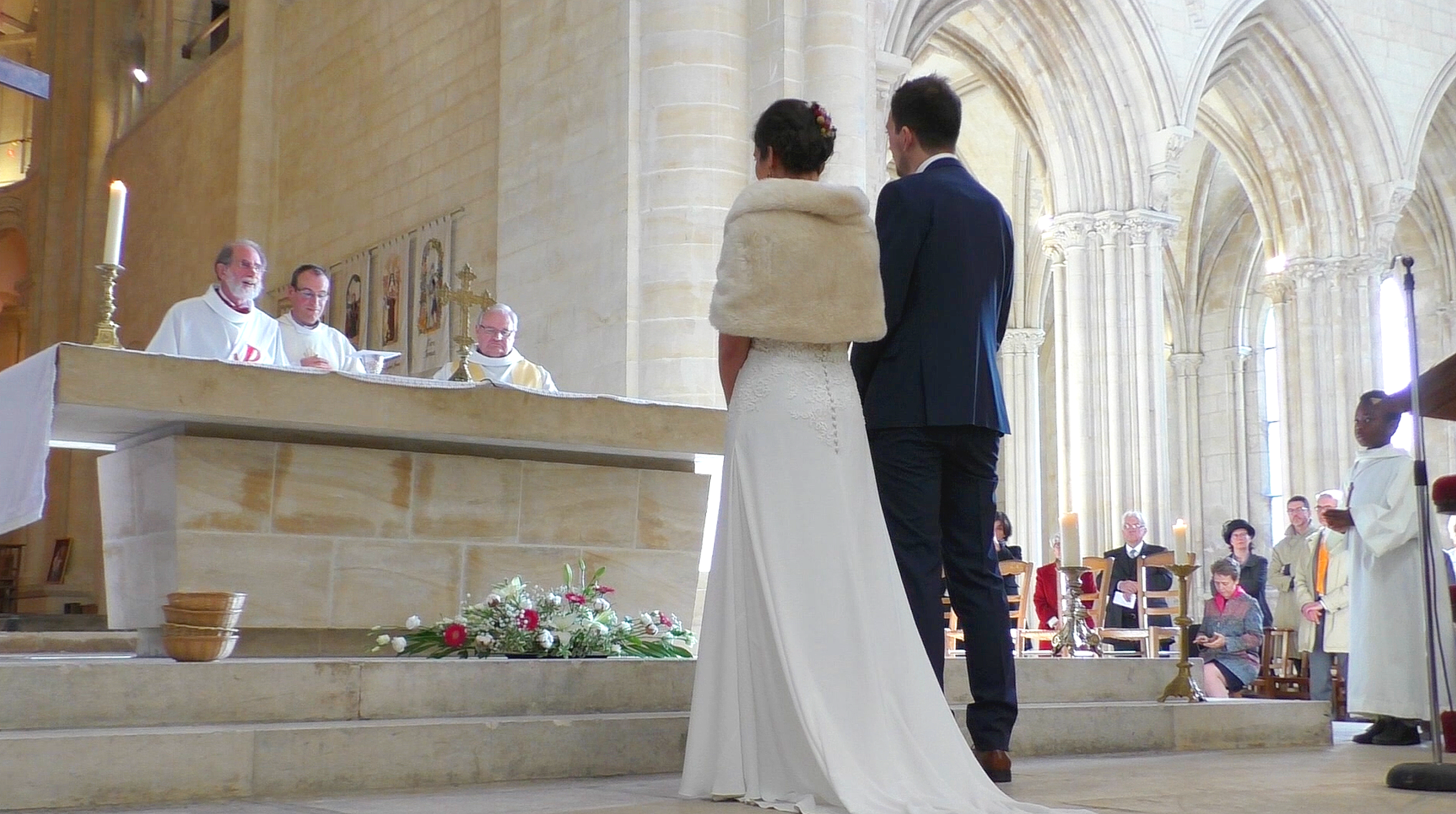 Un couple en tenue de marié devant un prêtre lors d'une cérémonie religieuse dans une église gothique. La mariée porte une robe blanche et une étole en fourrure, le marié porte un costume sombre. Des personnes assises en arrière-plan ainsi qu'un assistant en robe blanche sont visibles.