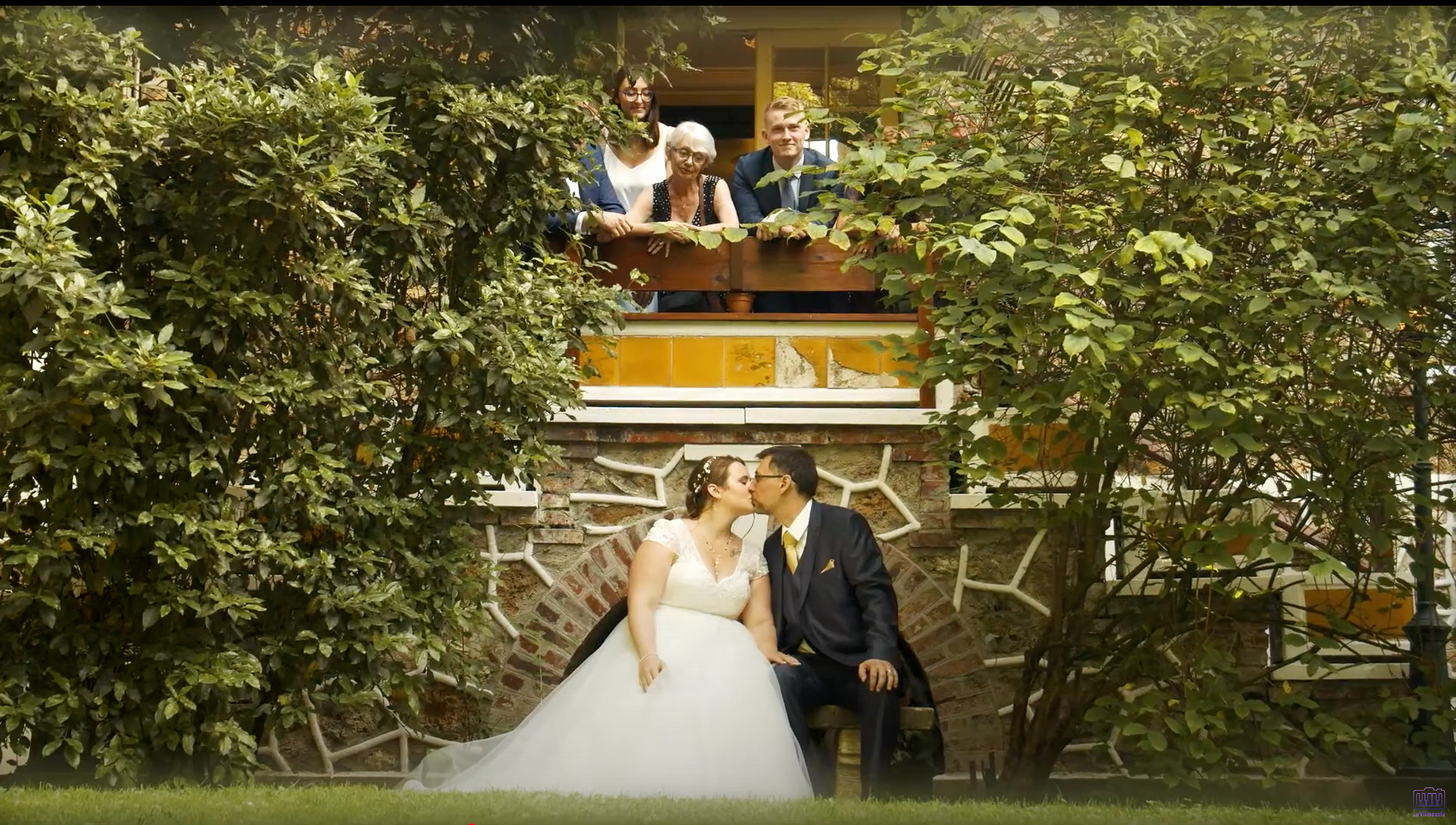 Un couple de mariés s'embrasse devant une maison en pierre avec des murs en briques et entourés de buissons verts, avec des invités regardant depuis le balcon.