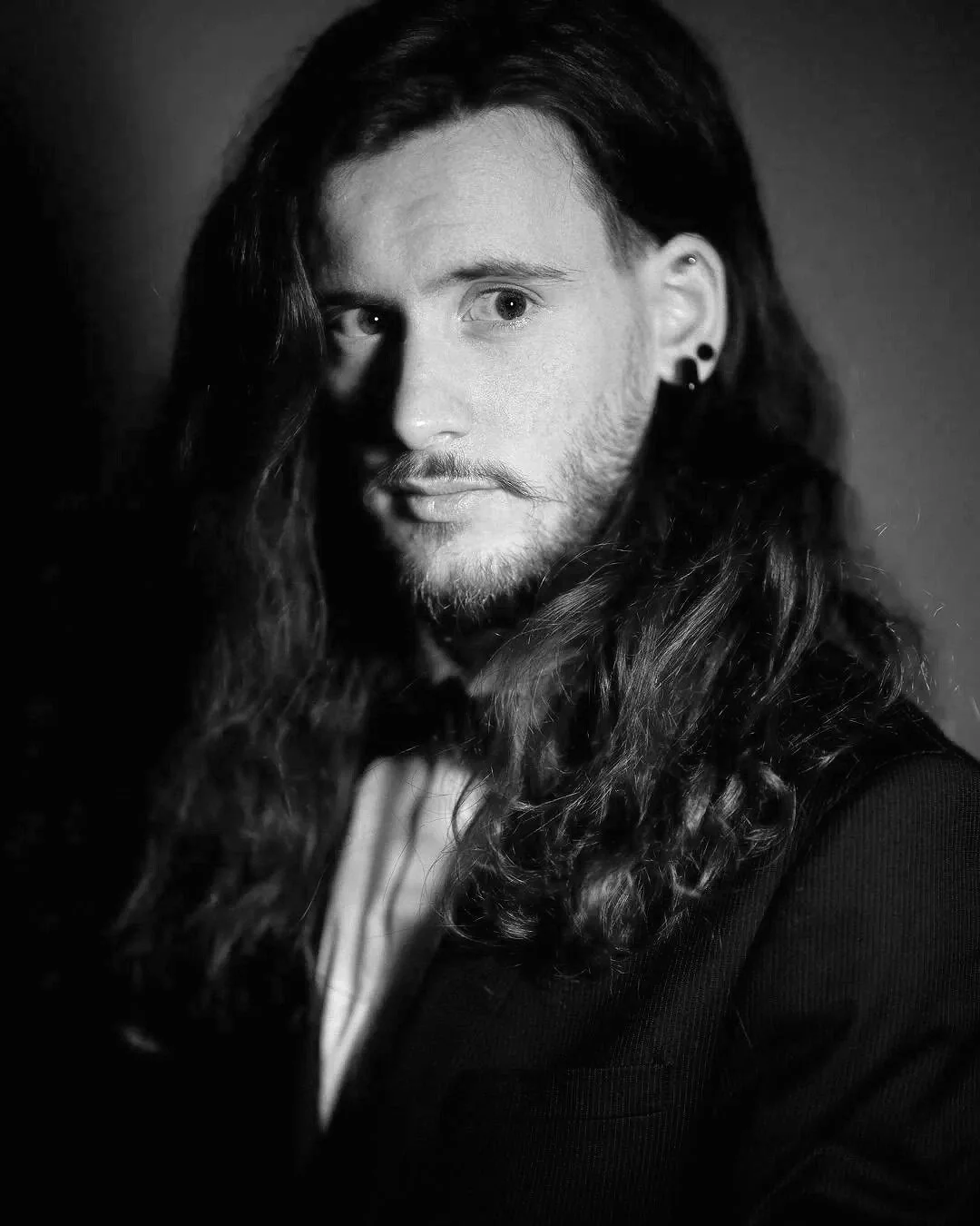 Portrait en noir et blanc d'un homme avec de longs cheveux ondulés, une barbe et portant des boucles d'oreilles, regardant vers l'objectif.