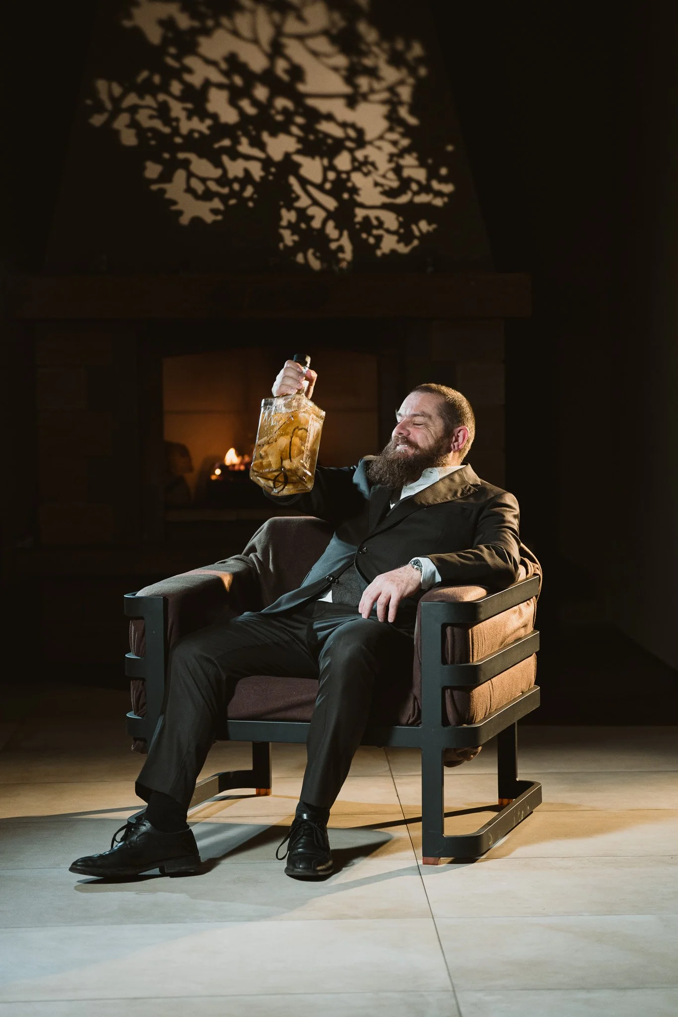 Un homme habillé en costume noir, assis dans un fauteuil, tenant une bouteille de liquor avec des citrons, dans une pièce sombre avec un feu au fond. Illustration artistique et élégante.