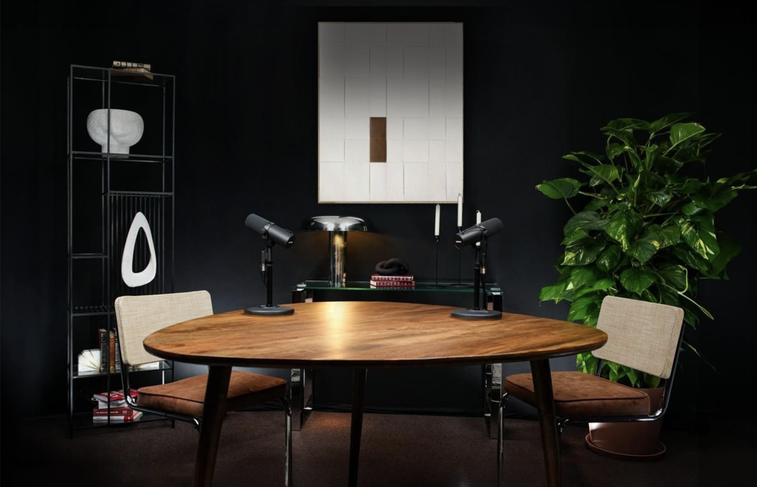 Studio de tournage podcast moderne avec une table en bois, six chaises en tissu beige, un tableau mural blanc, une étagère noire avec des livres et une œuvre d'art, une grande plante verte, deux lampes de bureau sur la table.