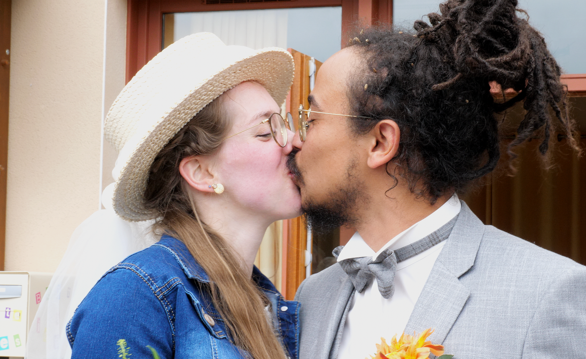 Deux personnes s'embrassent devant une porte en bois, une femme portant un chapeau en paille et des lunettes, un homme avec des dreadlocks, un costume gris et une cravate grise, tenant un bouquet de fleurs orange.
