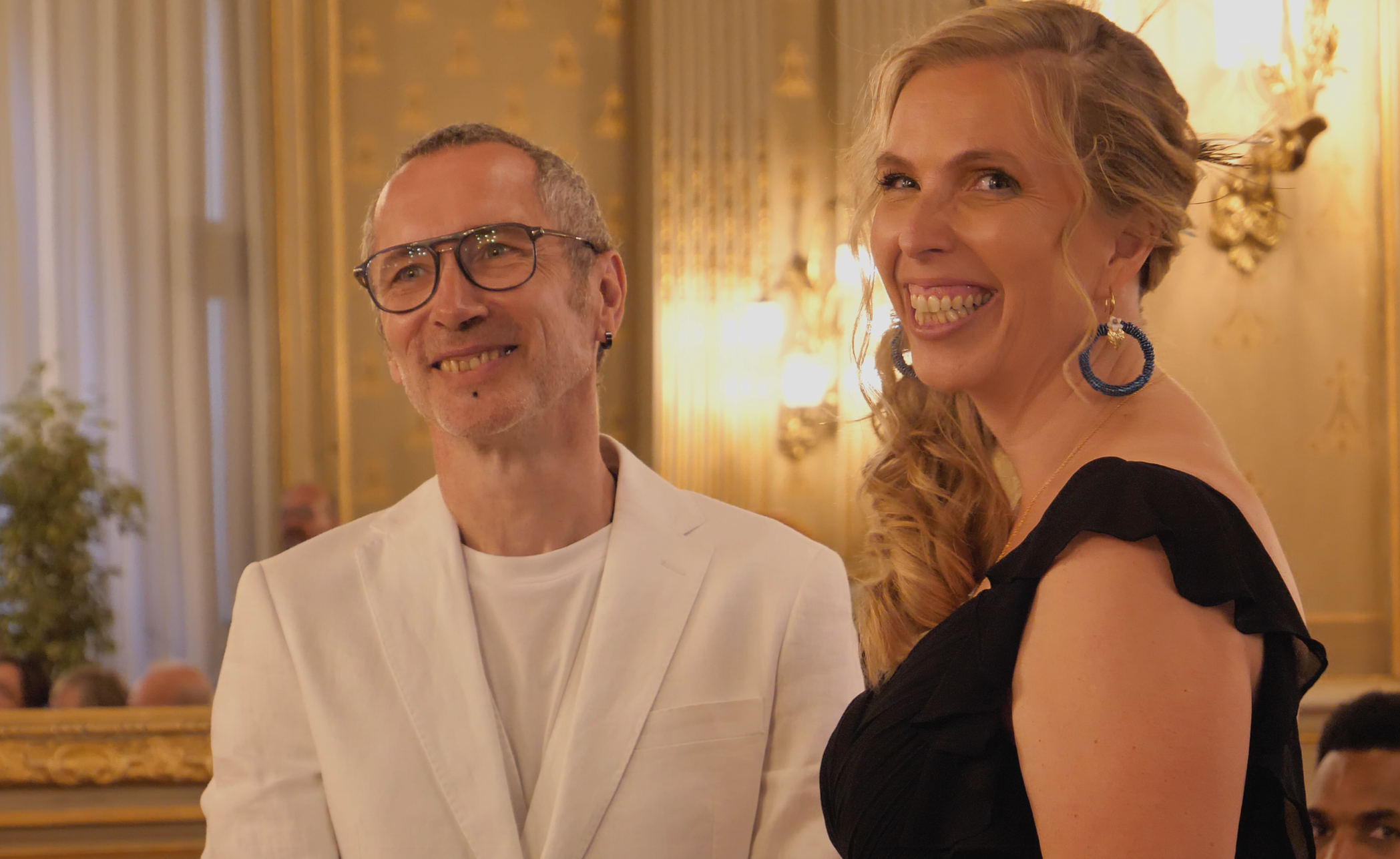 Un homme et une femme souriant lors d'un événement dans une salle ornée de dorures et de lumières chaleureuses.