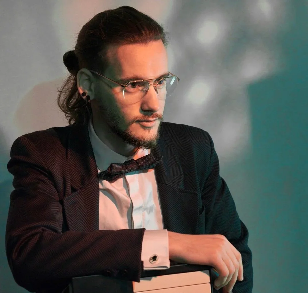 Jeune homme portant un costume noir avec un nœud papillon, lunettes et cheveux longs attachés, posant avec un ordinateur portable.