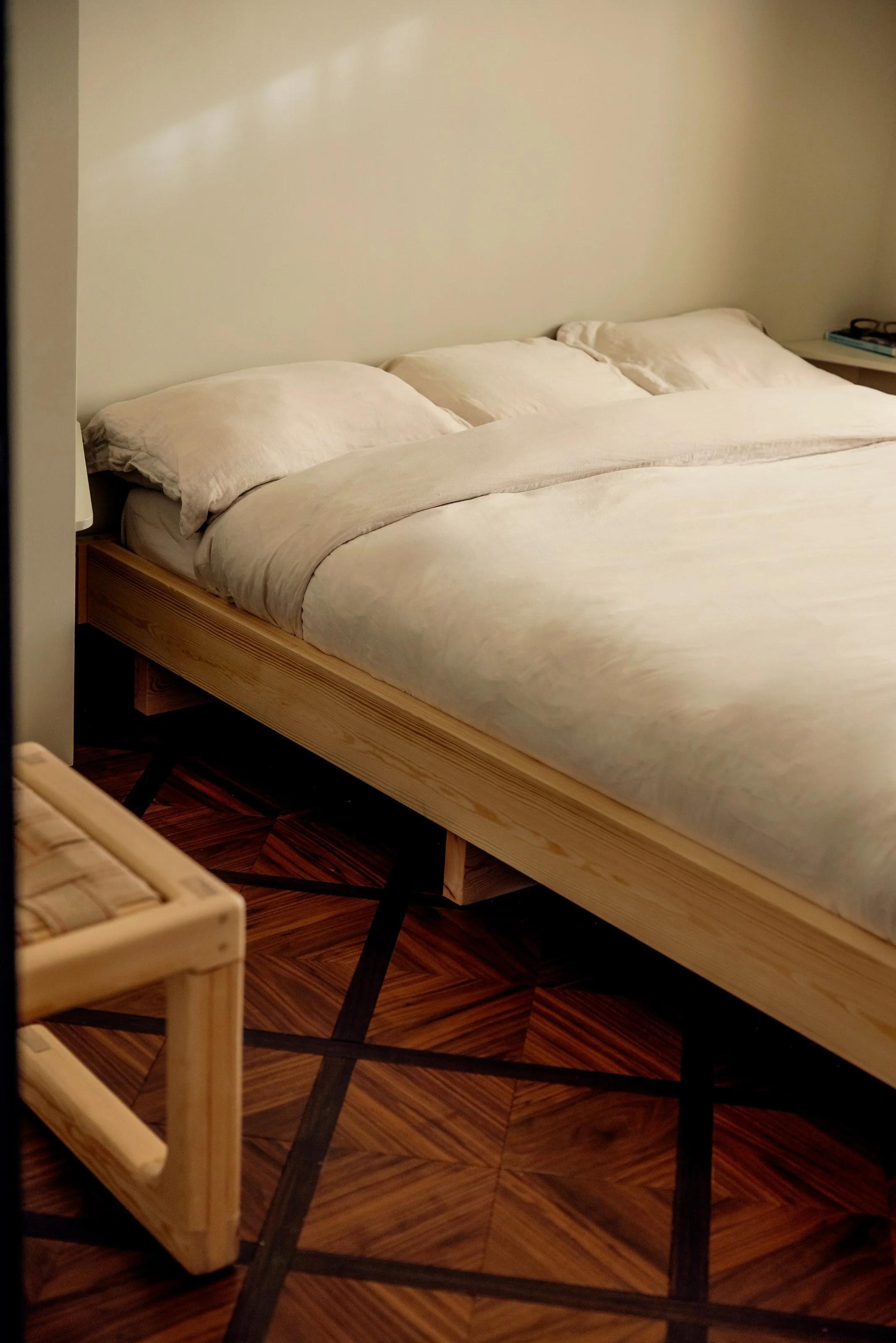 A bed with a beige or off-white duvet covers and matching pillows, set against a plain beige wall. To the right of the bed, there is a small side table with a pair of glasses and a few books on it. The floor is made of wood with a geometric pattern.