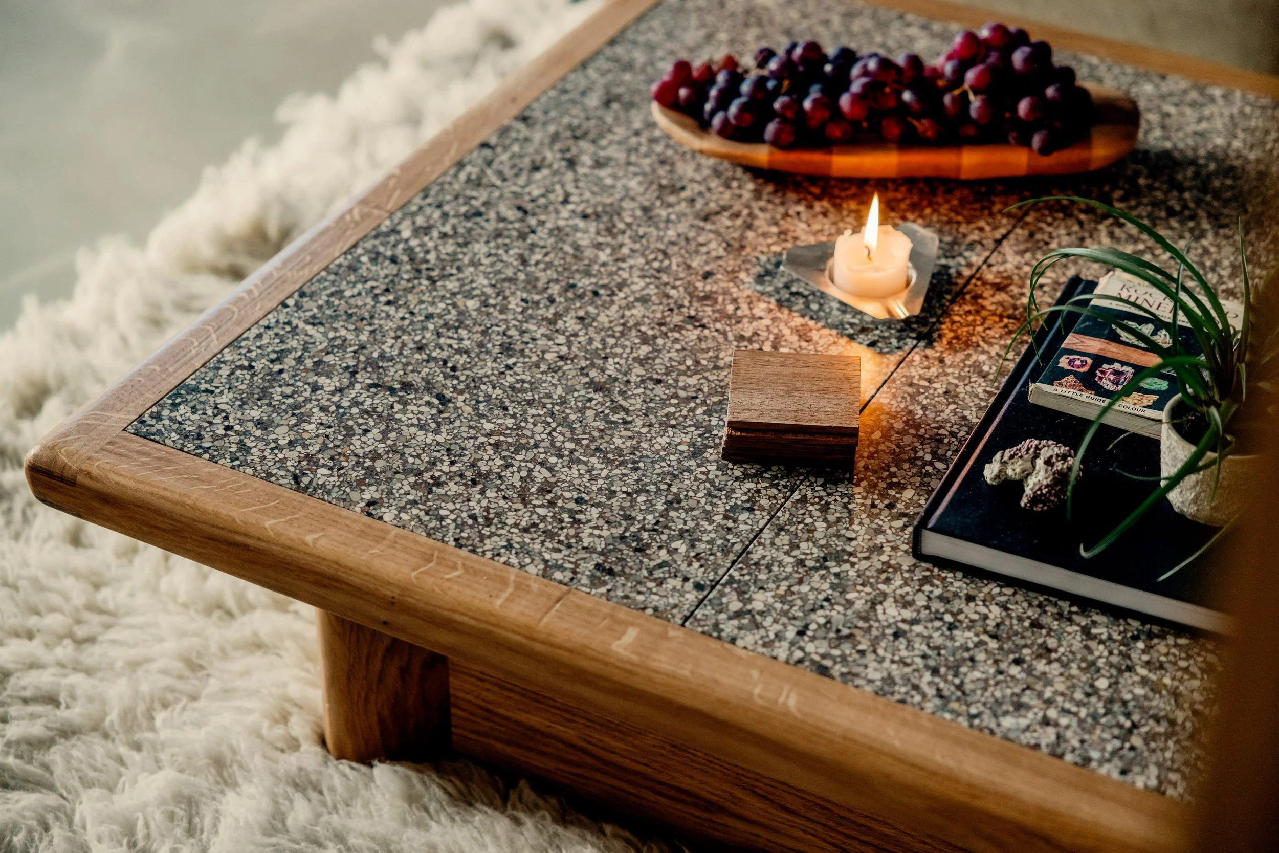 A tabletop with a speckled surface, a lit candle, a small wooden block, a tray with a plant, a book, and a wooden bowl filled with grapes, next to a soft, white shaggy rug.