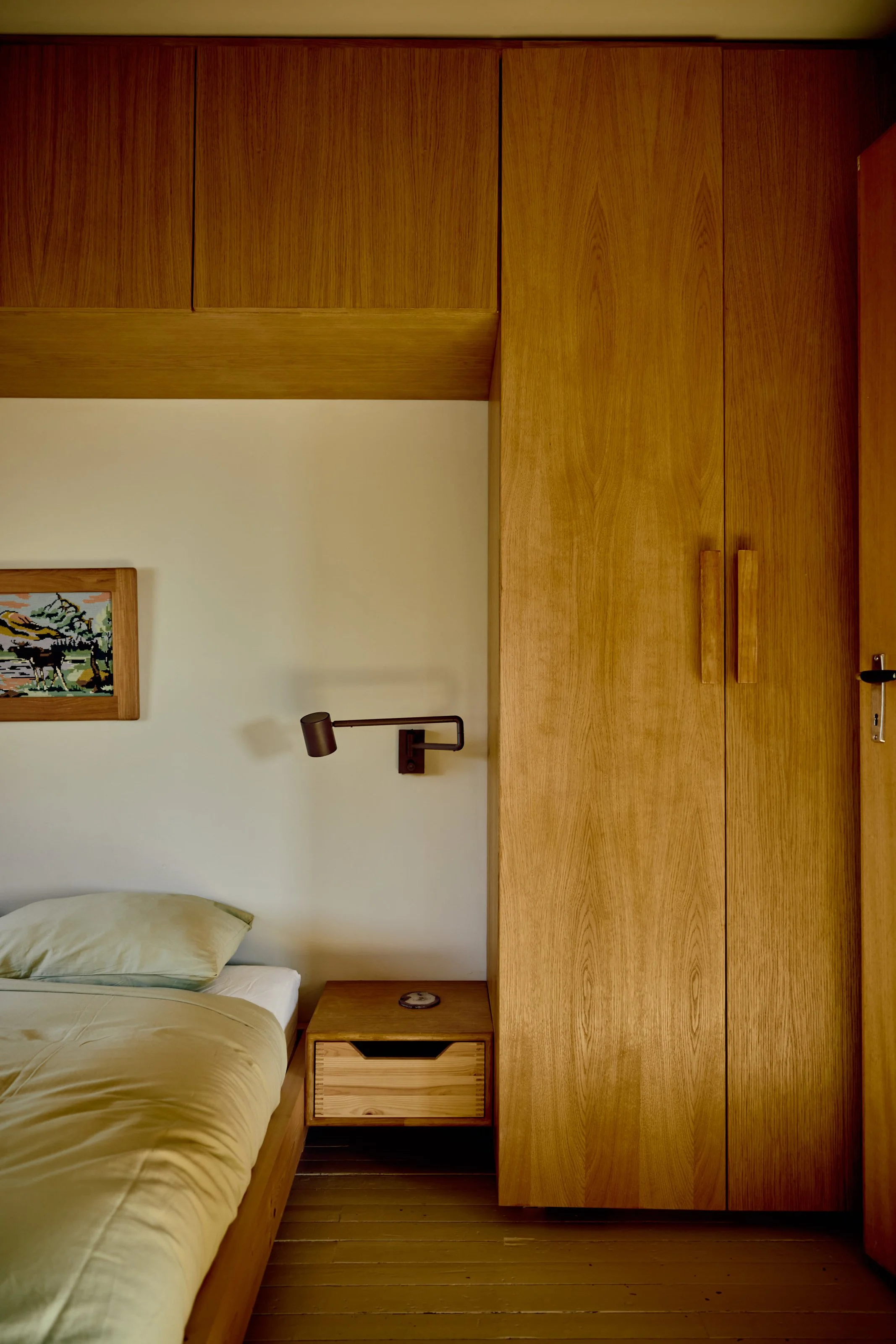 A bedroom with a wooden headboard, nightstand, and wardrobe, a bed with yellow bedding, a framed artwork on the wall, and a wall-mounted lamp and clock.