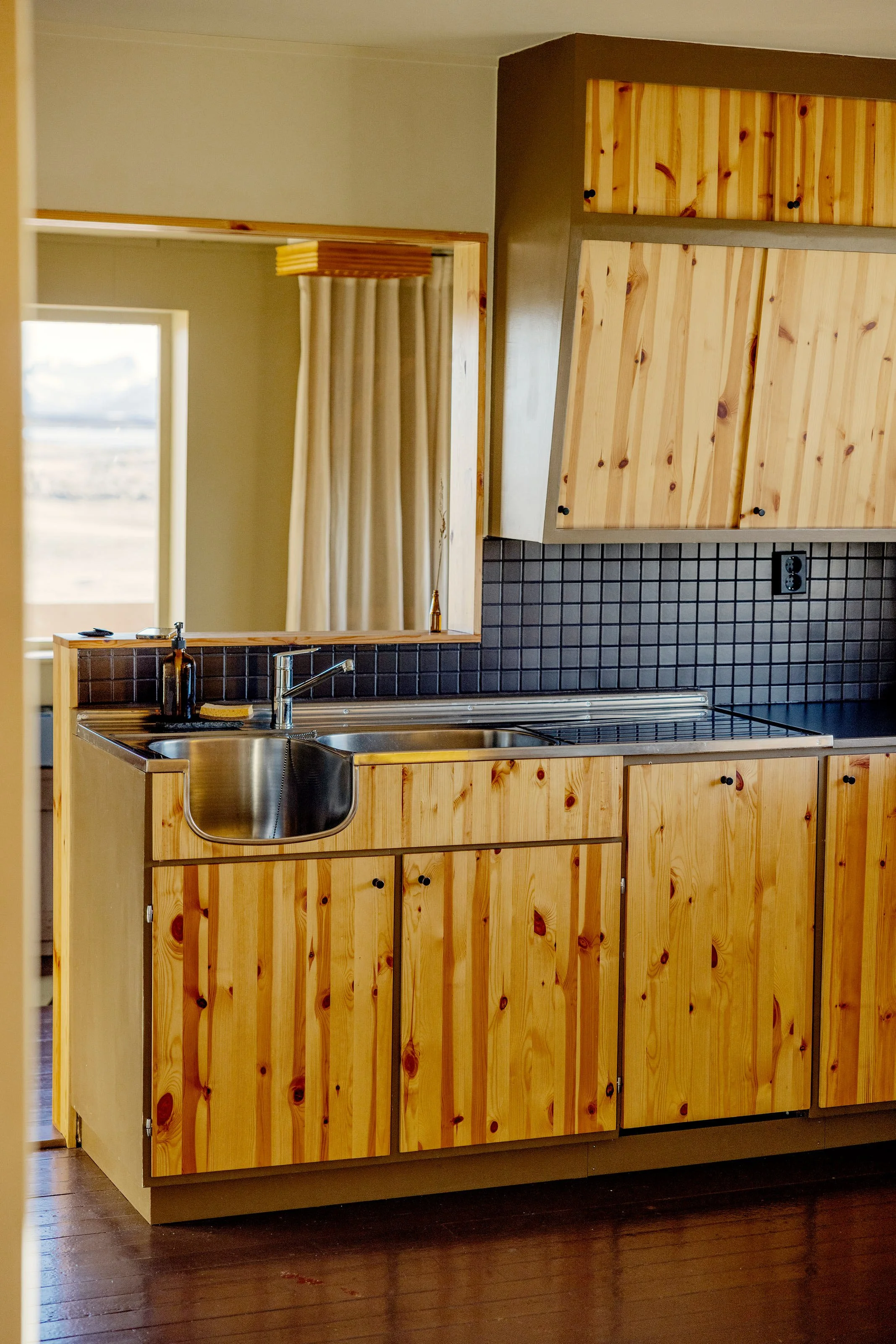 View of a wooden kitchen cabinet with black knobs, a small stainless steel sink, and a kitchen counter with a black tiled backsplash. A mirror on the wall reflects a window with beige curtains, and part of the room with light-colored walls and wood f