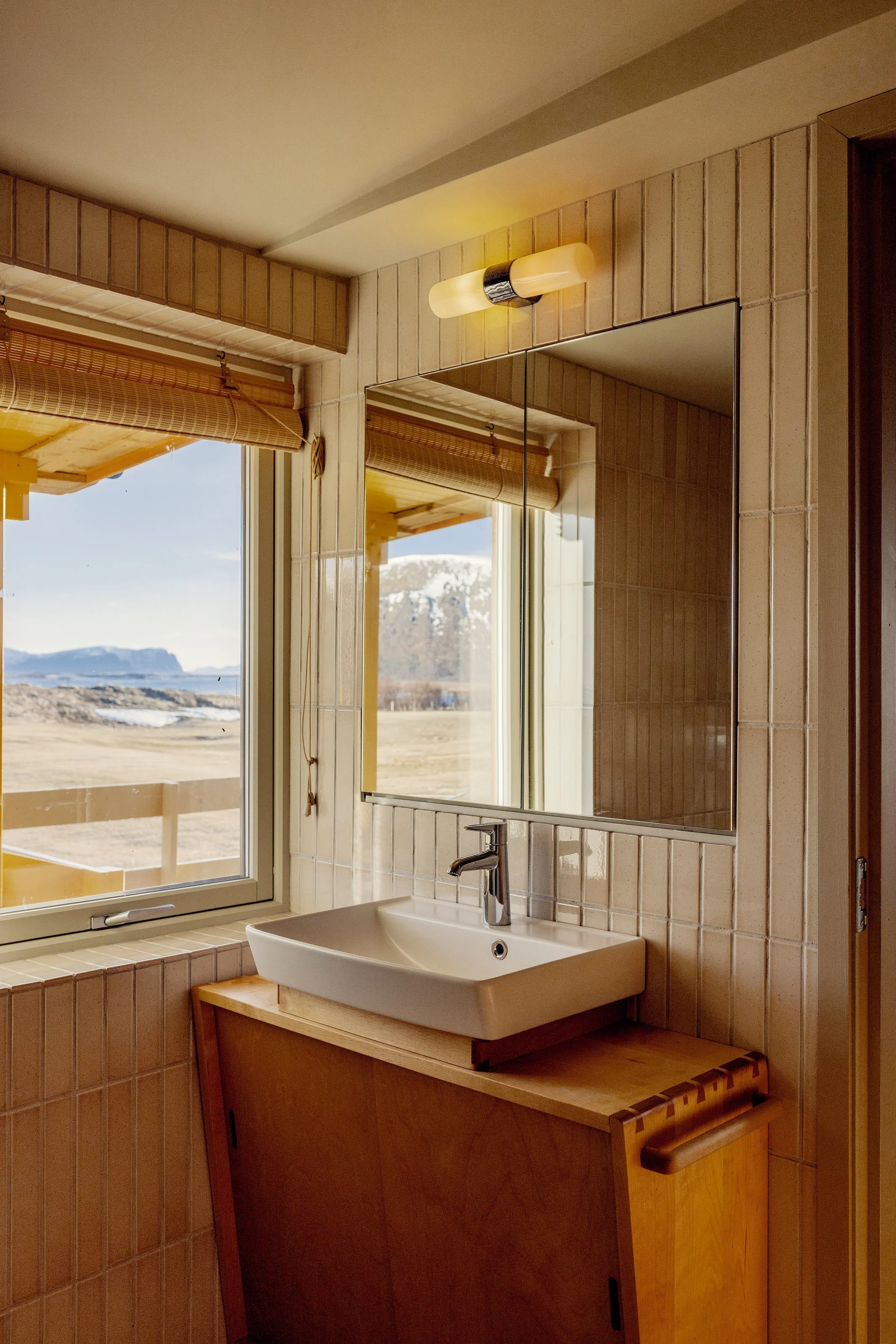 A bathroom with a white sink on a wooden stand, beige tile walls, a large mirror, a window with a bamboo roll-up blind, and a view of snow-capped mountains outside.
