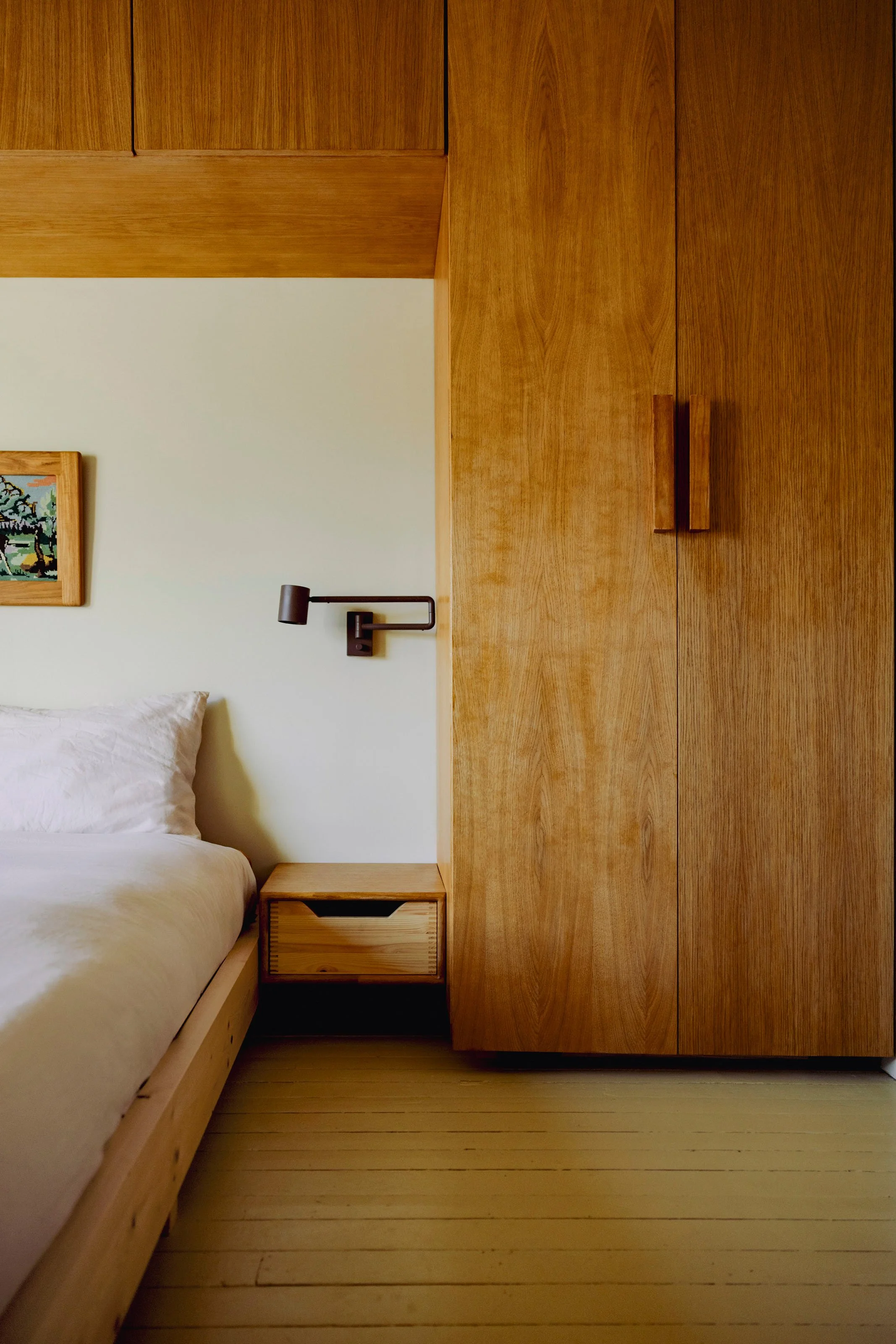 A bedroom with a bed, a wooden nightstand, a wall-mounted black reading lamp, and a large wooden wardrobe. There is a small framed artwork on the wall near the bed.