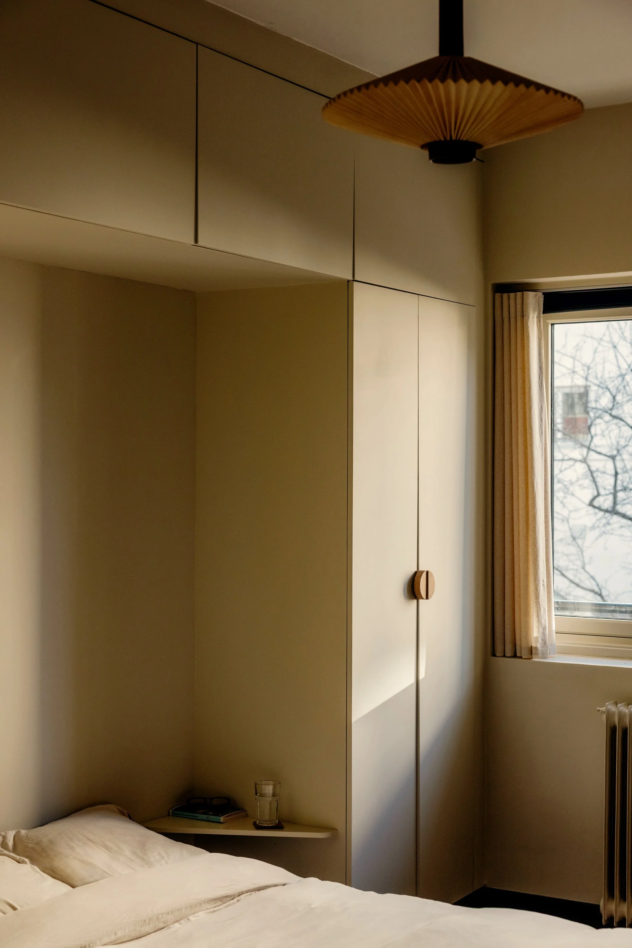 A cozy bedroom corner with a bed, a small side table holding a glass of water and books, beige built-in cabinets on the wall, and a window with cream curtains showing leafless trees outside.