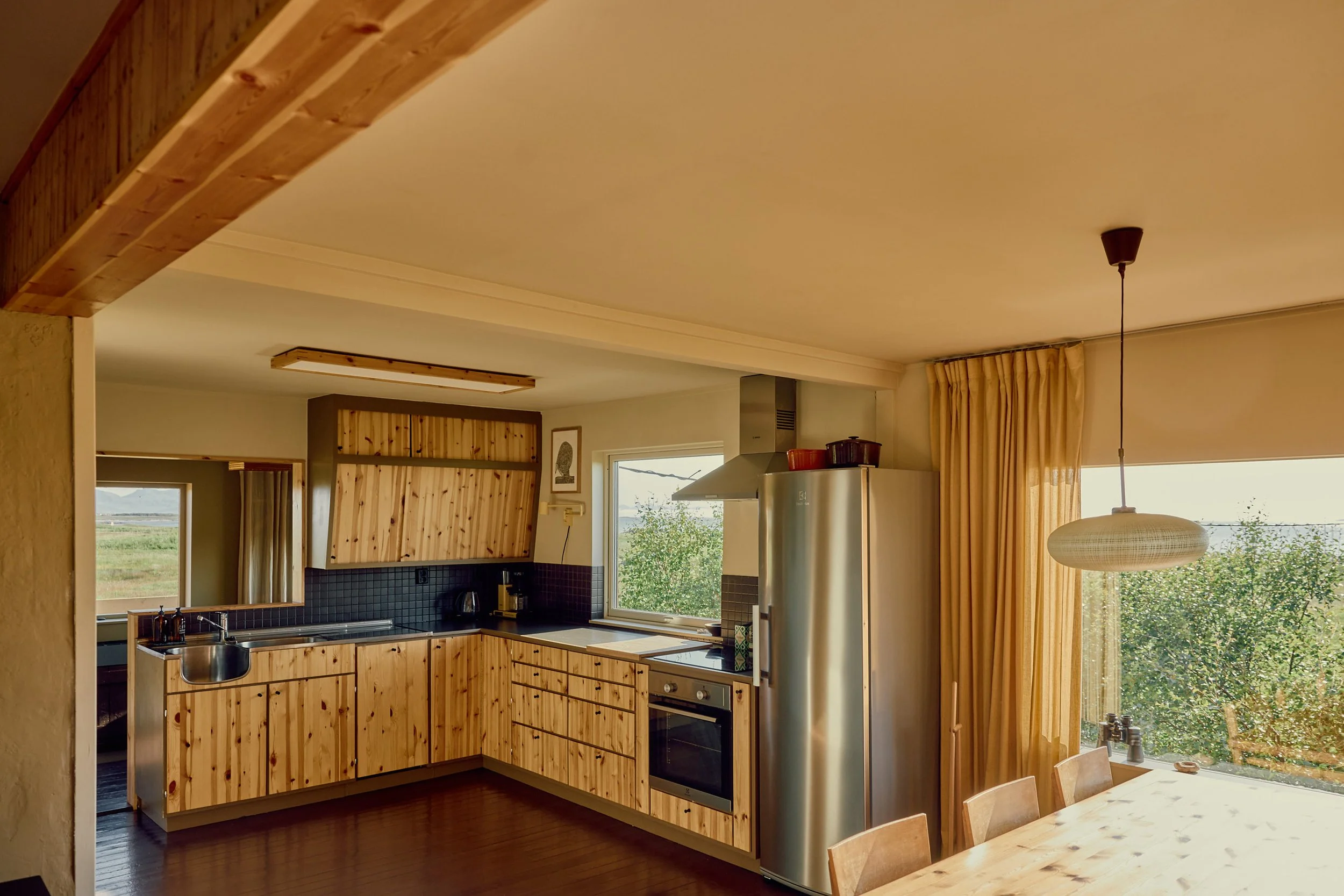 A kitchen with wood cabinets, black backsplash, and a stainless refrigerator near large windows with yellow curtains. There is a wooden dining table with chairs and a hanging light fixture.