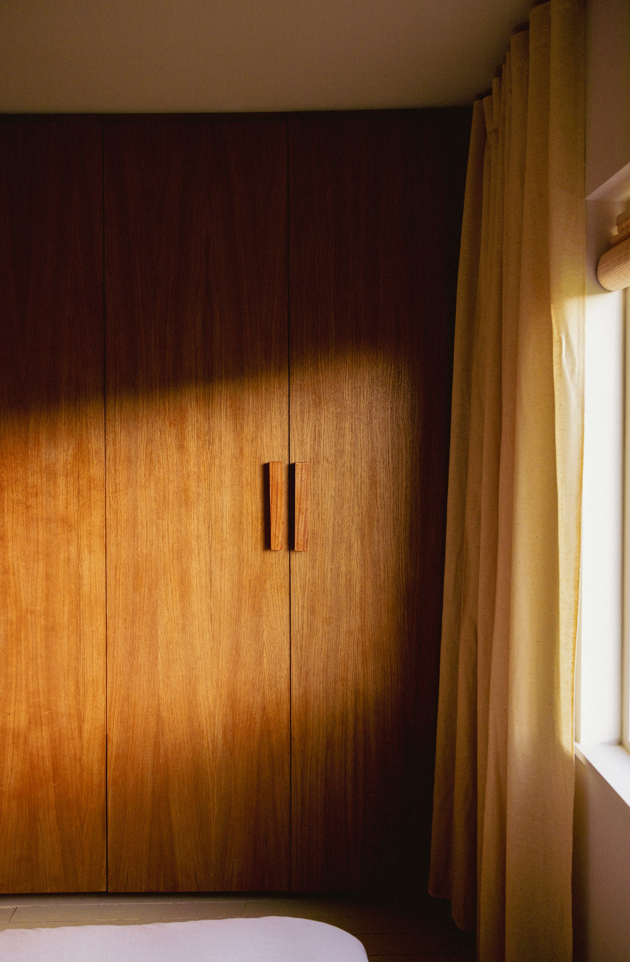 Wooden wardrobe with two doors and handles, beige curtains, and a window with sunlight in the room.
