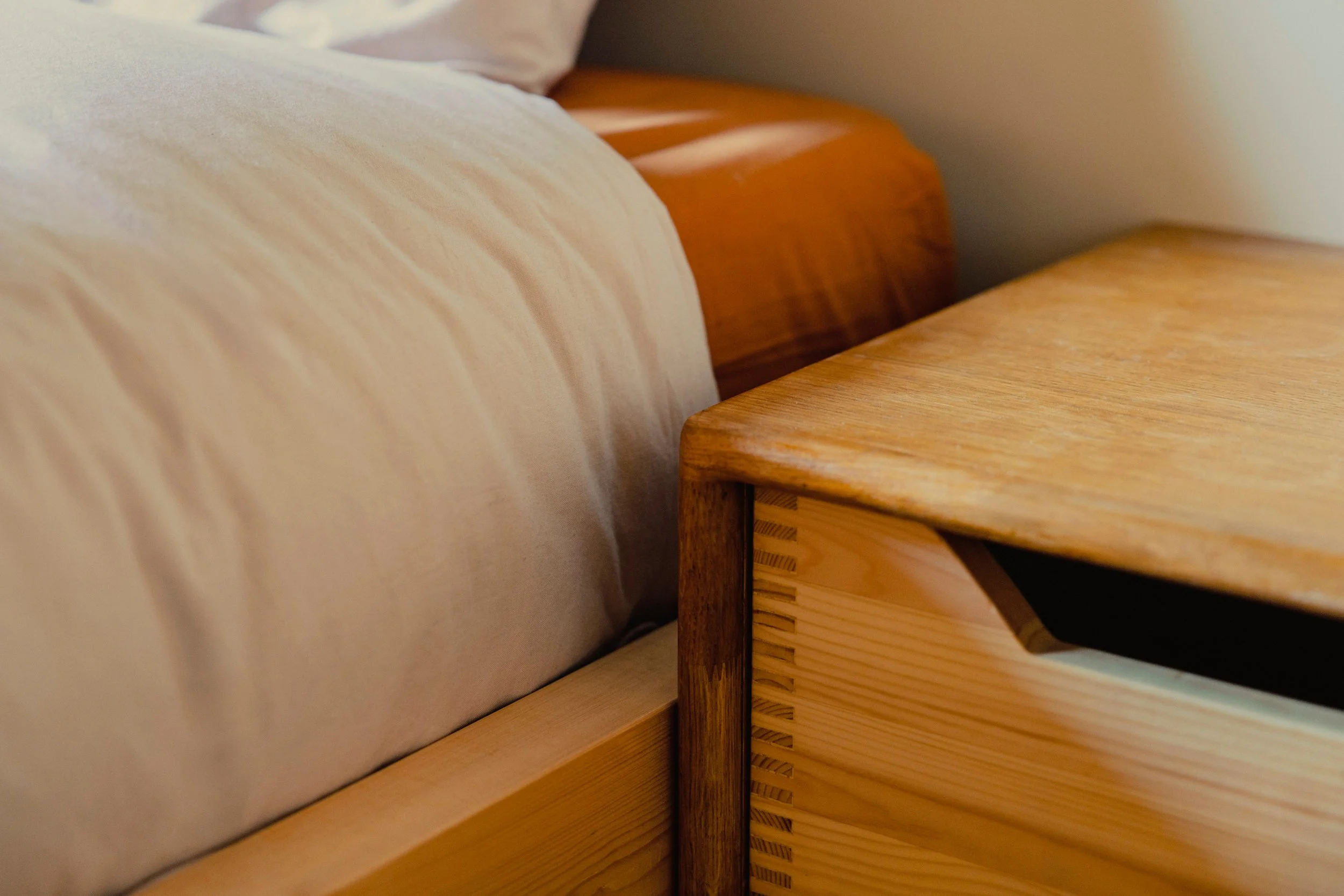 Close-up of wooden furniture pieces, including a bed frame and a nightstand, with a bed and a pillow in the background.