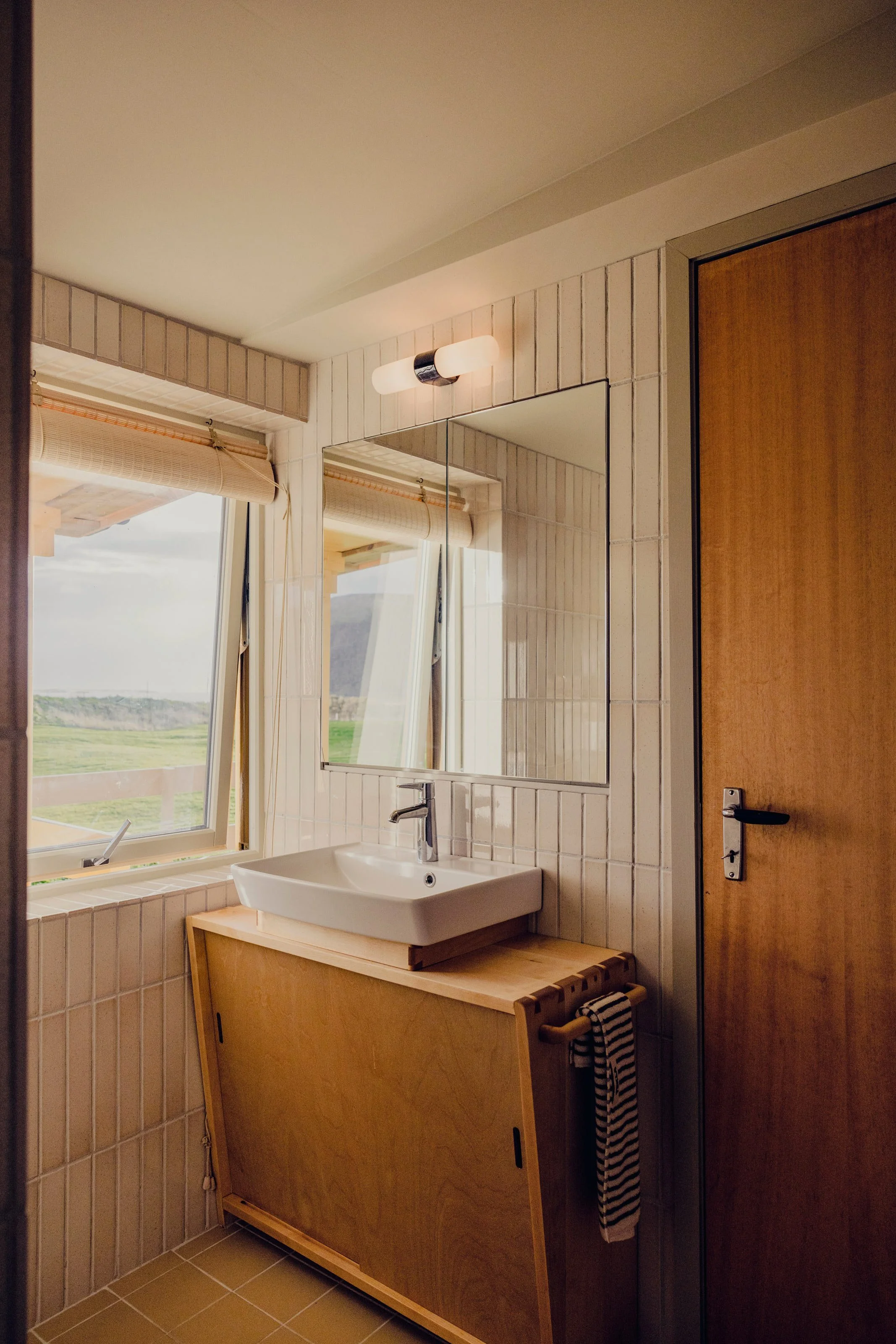 A bathroom with a window showing a landscape, a white sink with a wooden cabinet underneath, a mirror above the sink, a light fixture above the mirror, and a wooden door on the right side. There is a black and white striped towel hanging on a rack be
