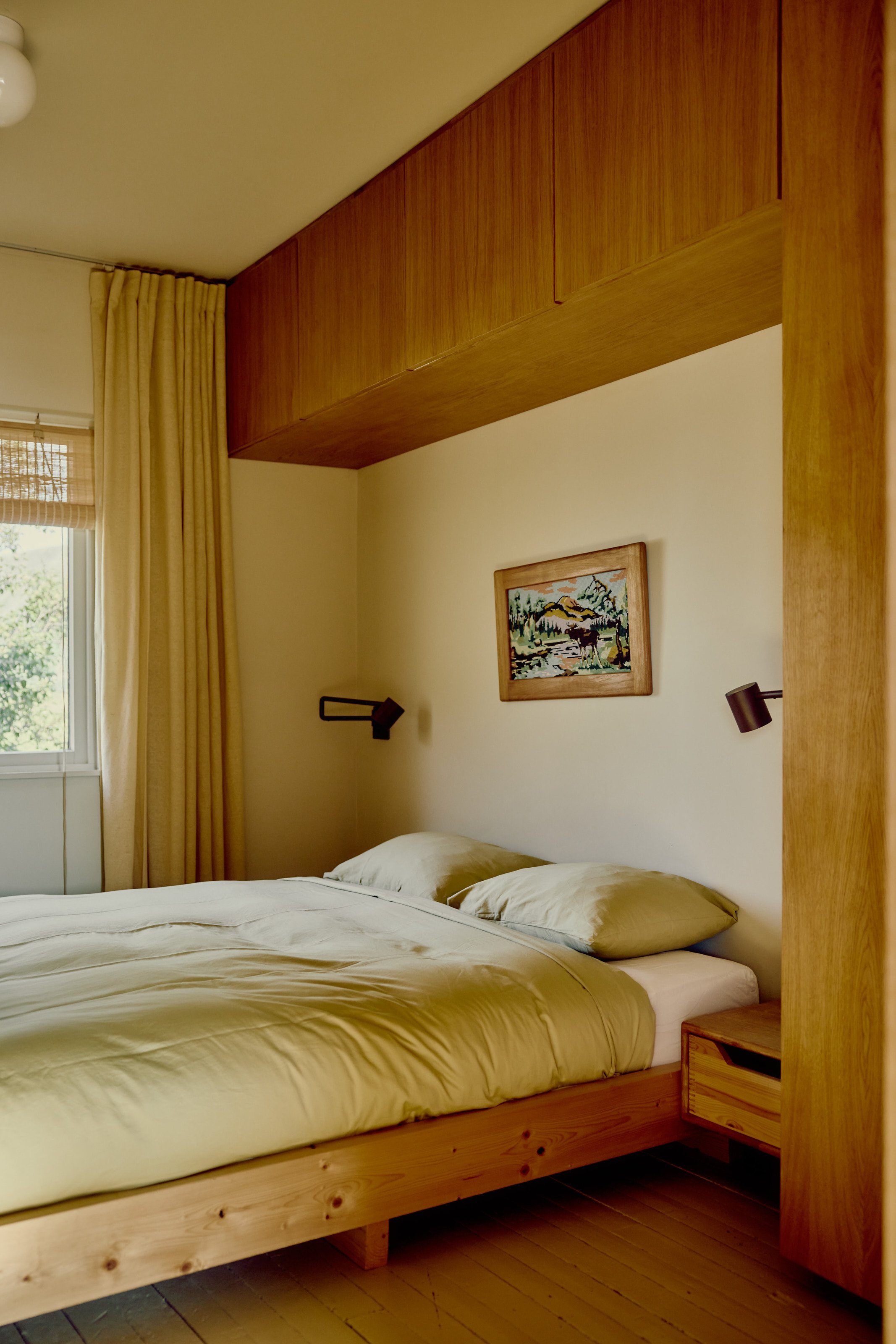 A cozy bedroom with a wooden bed frame, beige bedding, and matching pillows. There are light beige curtains by a window and a framed landscape painting on the wall above the bed. Wall-mounted black lamps are next to the painting.