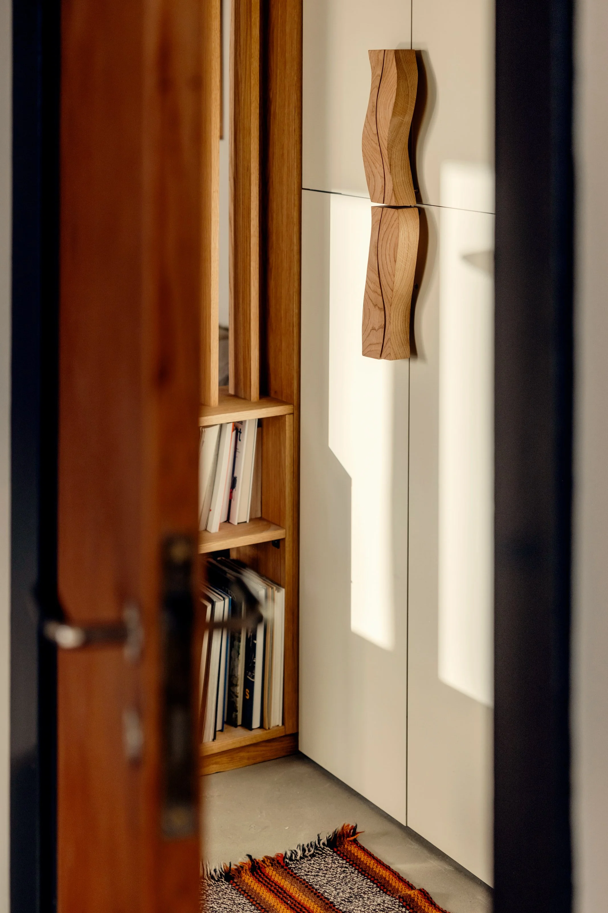 View of a room through a partially open door, showing a wooden bookshelf filled with books, a white cabinet with curved wooden handles, a colorful woven rug on the floor, and a small section of a wall with shadows cast by sunlight.