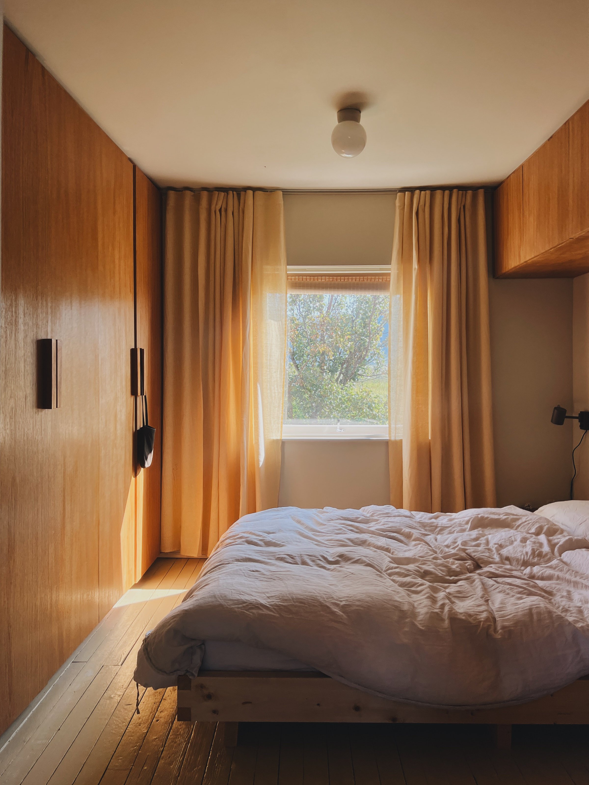 A bedroom with a wooden wardrobe, a bed with a crumpled blanket, and a window with beige curtains revealing trees outside.