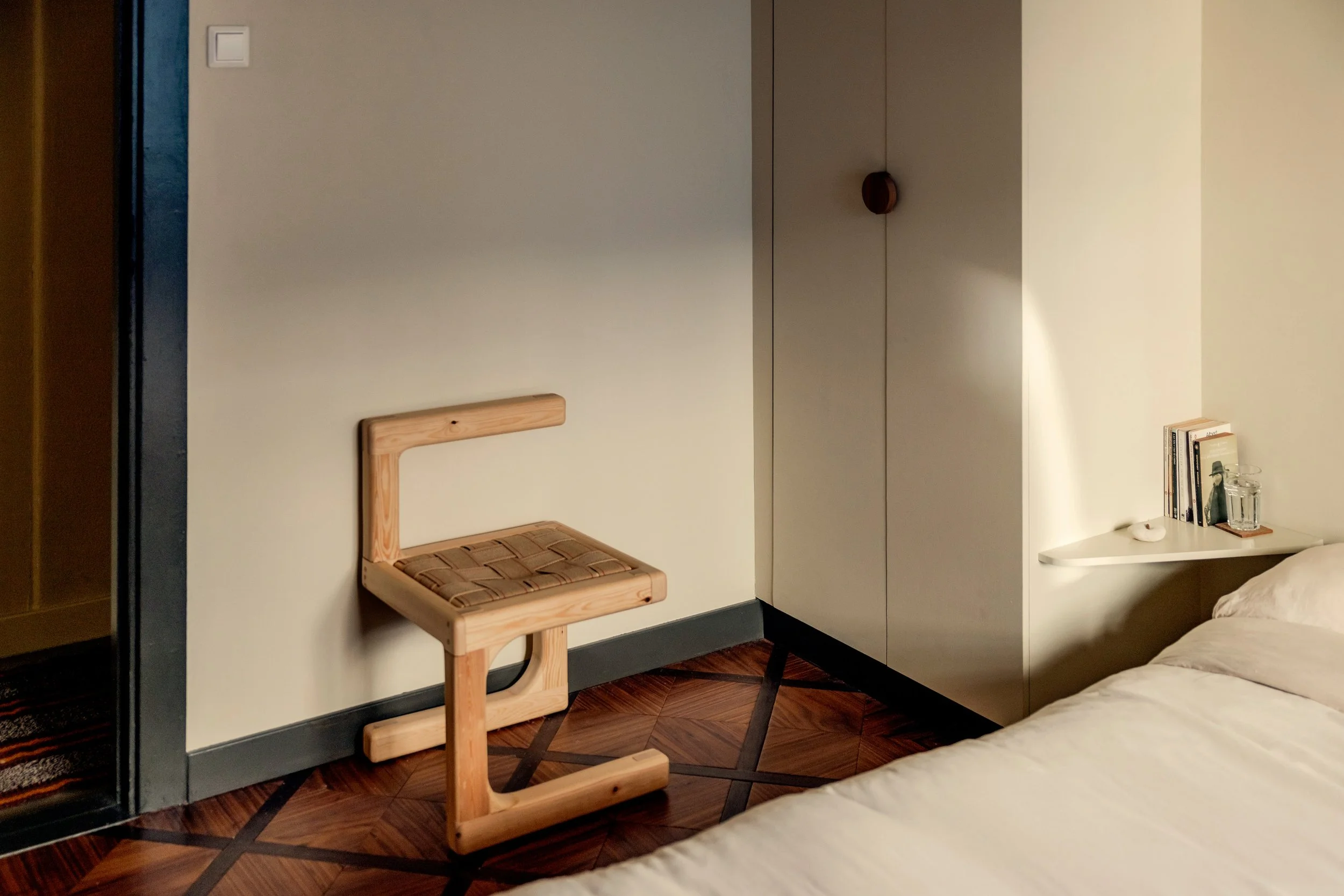 A bedroom corner with a bed, a small desk, and a hanging chair with a woven seat. The room has a wooden floor and minimal decorations.