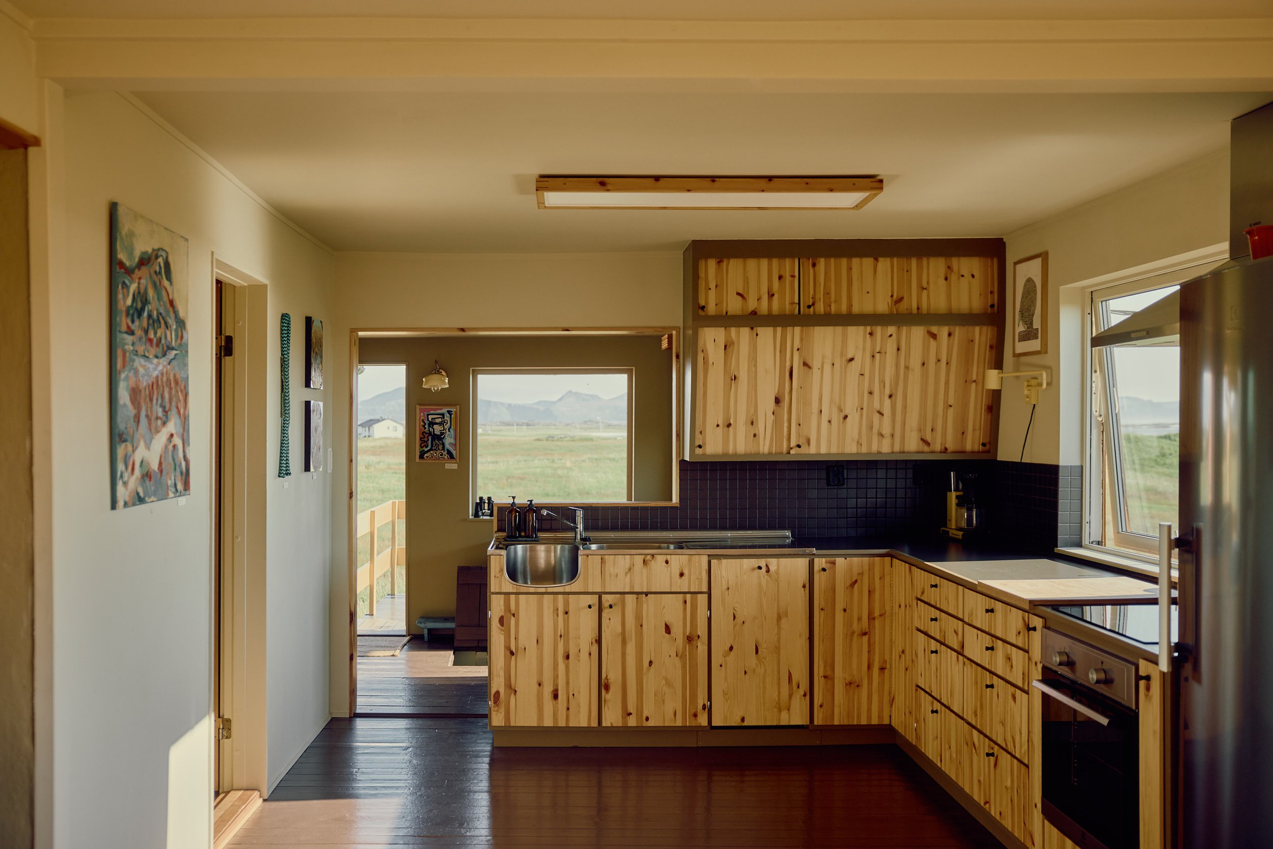 A kitchen with wooden cabinets, black countertop, stainless steel sink, and a window showing an outdoor view of mountains and fields.