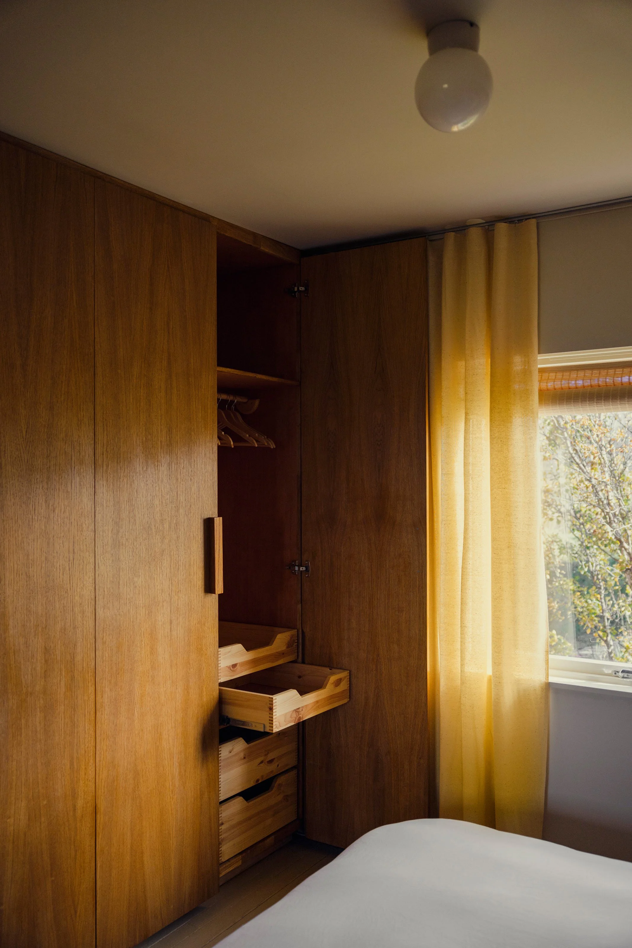 Interior of a bedroom with a wooden wardrobe, a window with yellow curtains, and a view of trees outside, with a wooden bed frame partially visible at the bottom.