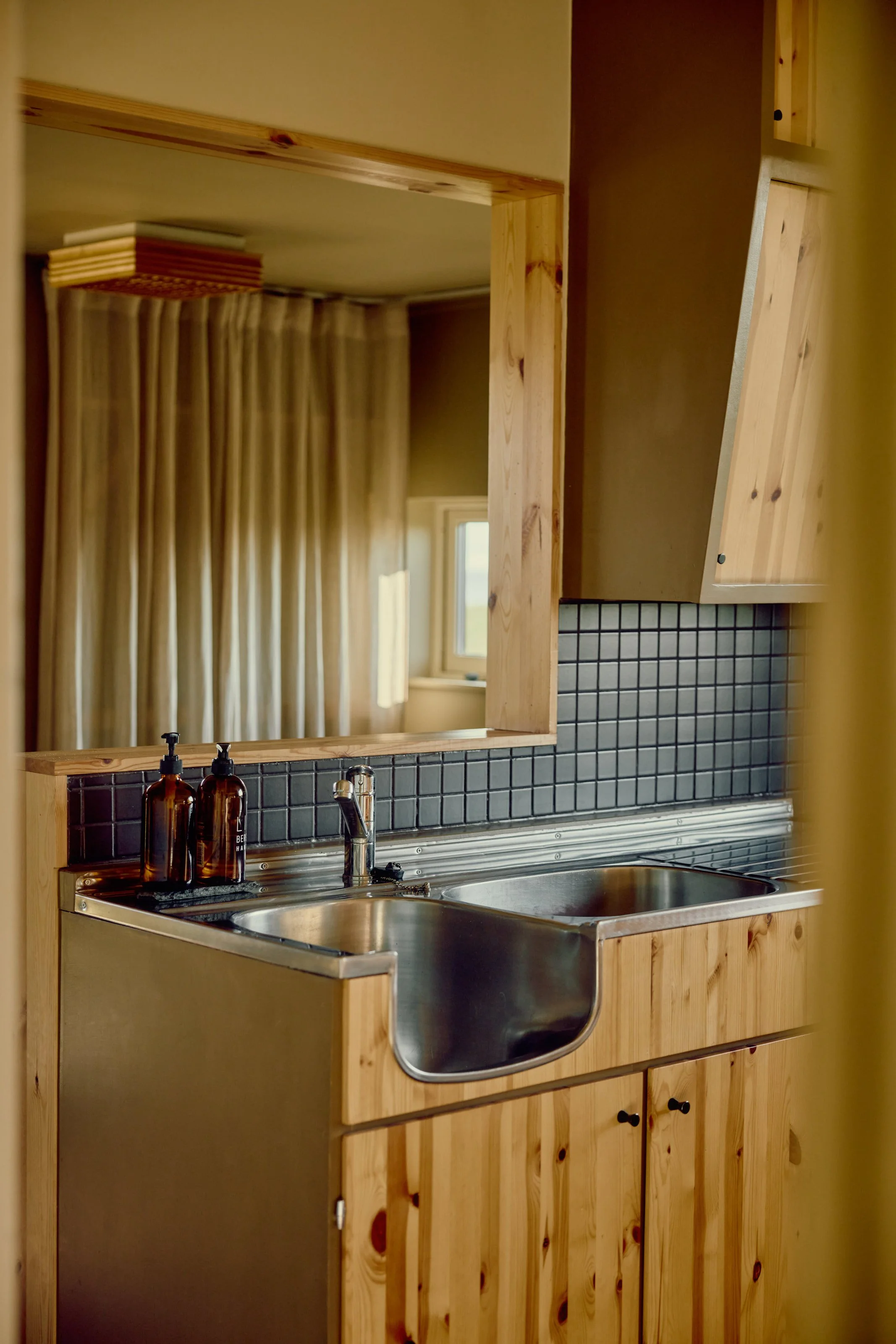 Wooden kitchen sink and countertop with black tile backsplash, stainless steel sink, and brown soap bottles, with a window and curtains in the background.