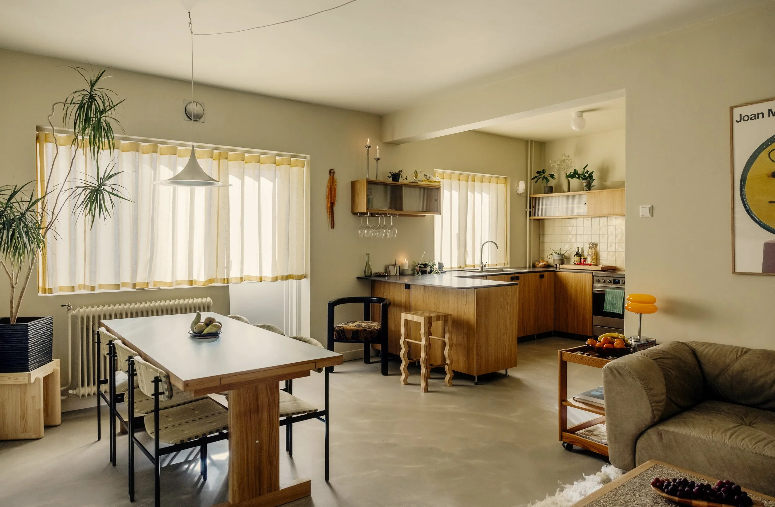 1940s Scandinavian apartment with concrete floor and oak kitchen. Warm light filtering through yellow waveband curtains. Shaker style dining table with blue formica table top. Italian 70s modular sofa. The space is featuring natural light and plants.