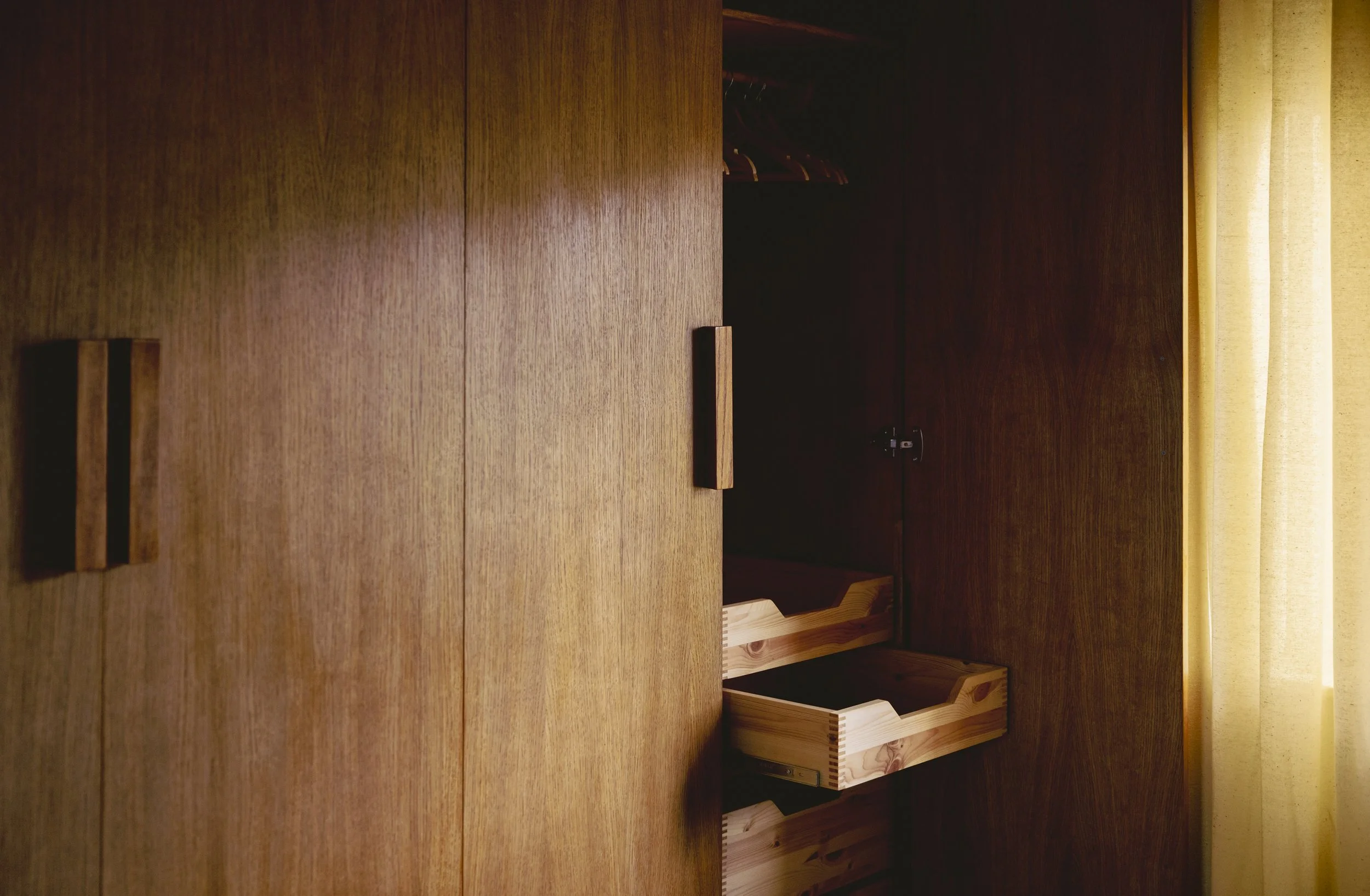 Retro oak Scandinavian built in wardrobe. Open wooden closet with hanging space and empty shelves, with sunlight coming through a yellow curtain.