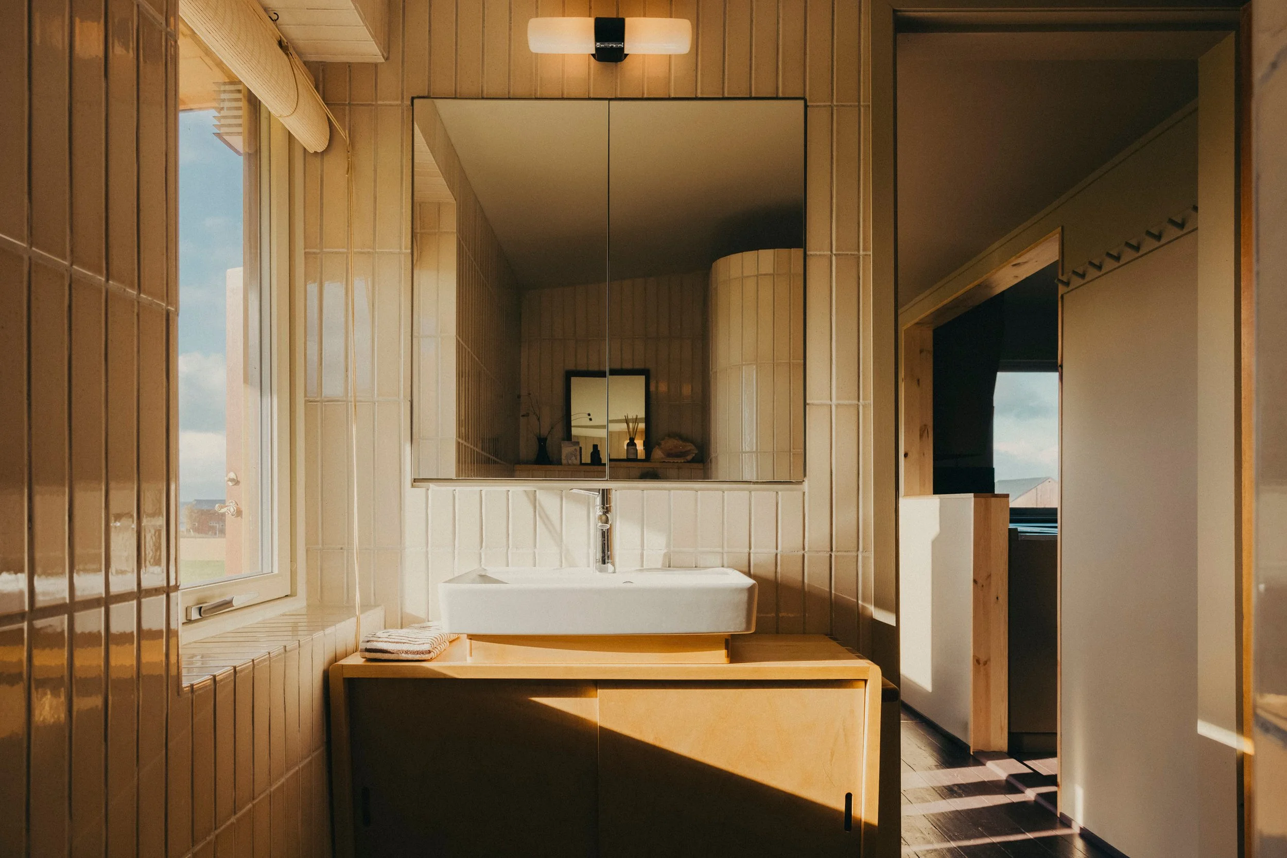 A bathroom with beige tiled walls and a large mirror above the sink. There is a window to the left with a view of a cloudy sky. A towel is placed on a wooden table below the sink. The bathroom is lit by a modern wall-mounted light fixture above the m