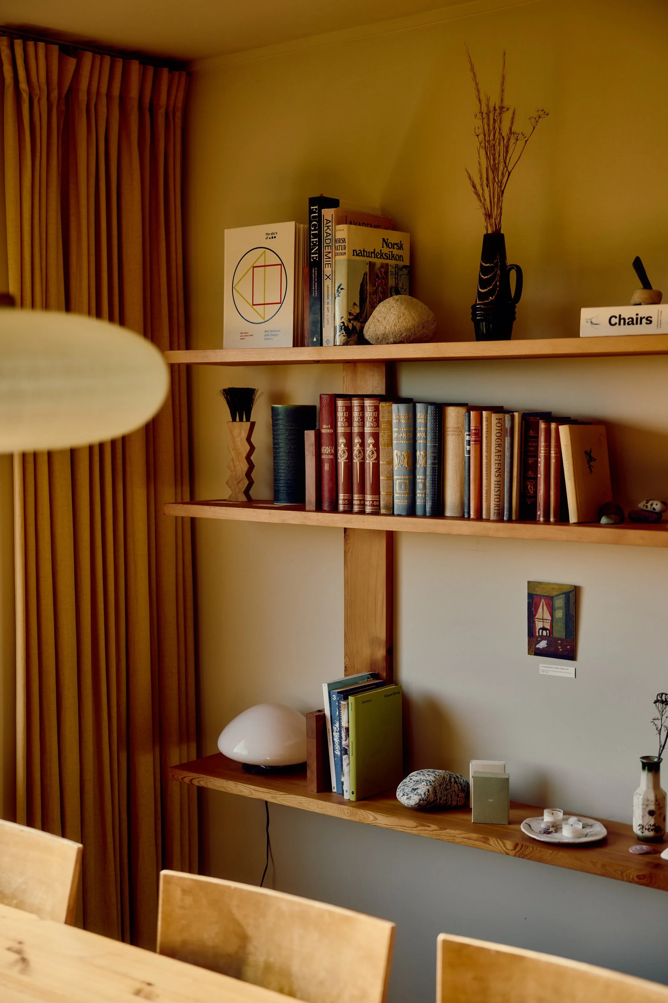 Wooden bookshelves with various books, decorative stones, a black vase with dried branches, a white table lamp, a small piece of art on the wall, a table with chairs in the foreground.