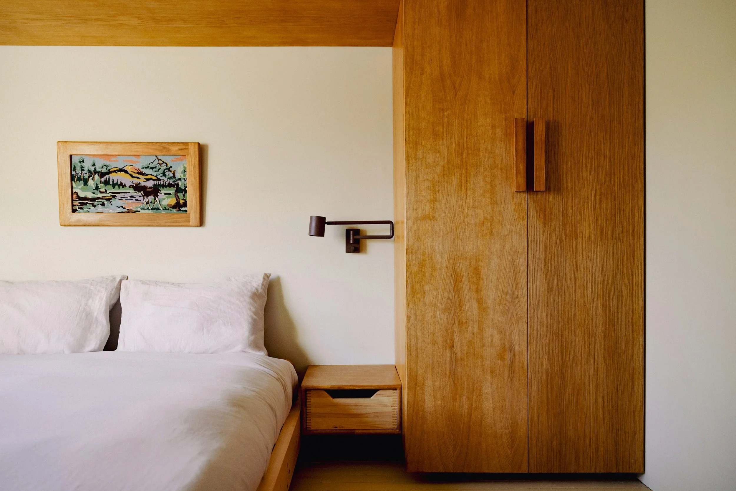 Close-up view of a bedroom with white bed linens, two white pillows, a wooden headboard, a small bedside table, and a wooden wardrobe in the background. A framed landscape painting featuring mountains, trees, and a river hangs on the wall above the b