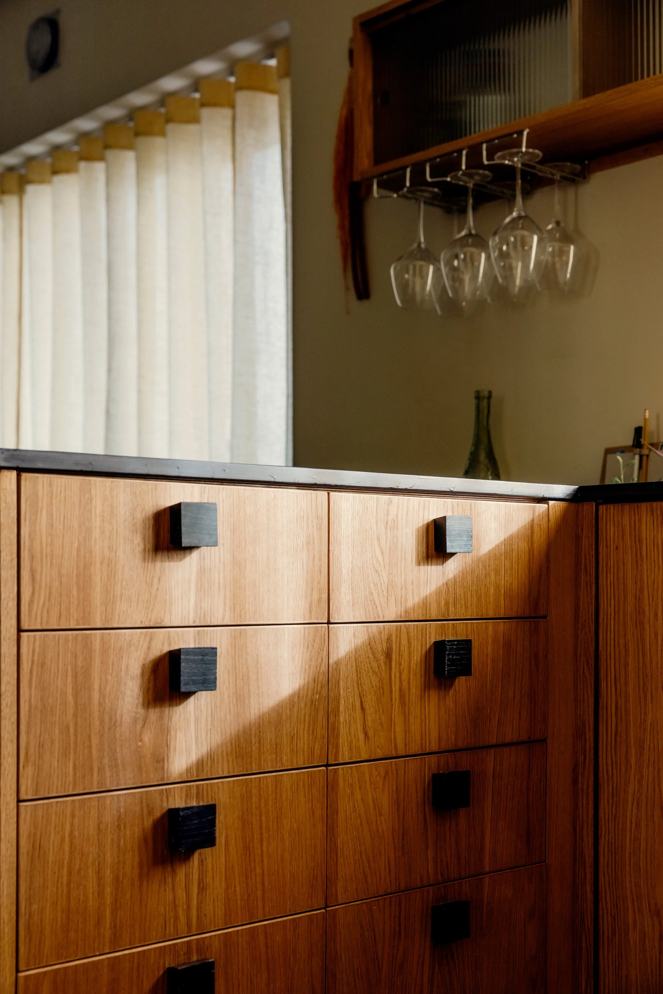 View of a wooden dresser with black square handles, a countertop, a decorative vase, and hanging wine glasses stored in a wooden rack, with a beige curtain and a wall-mounted clock in the background.