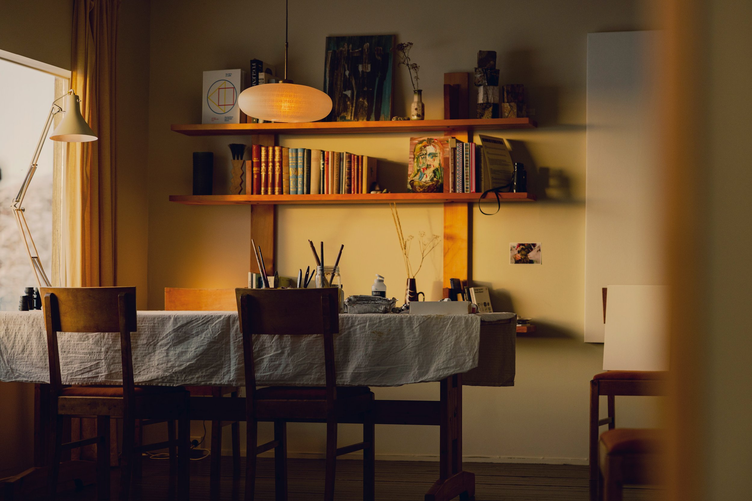 A cozy dining room with a table covered in a white tablecloth, surrounded by wooden chairs. A bookshelf mounted on the wall holds books and decorative items, illuminated by a hanging pendant light. A window with golden curtains lets in natural light 